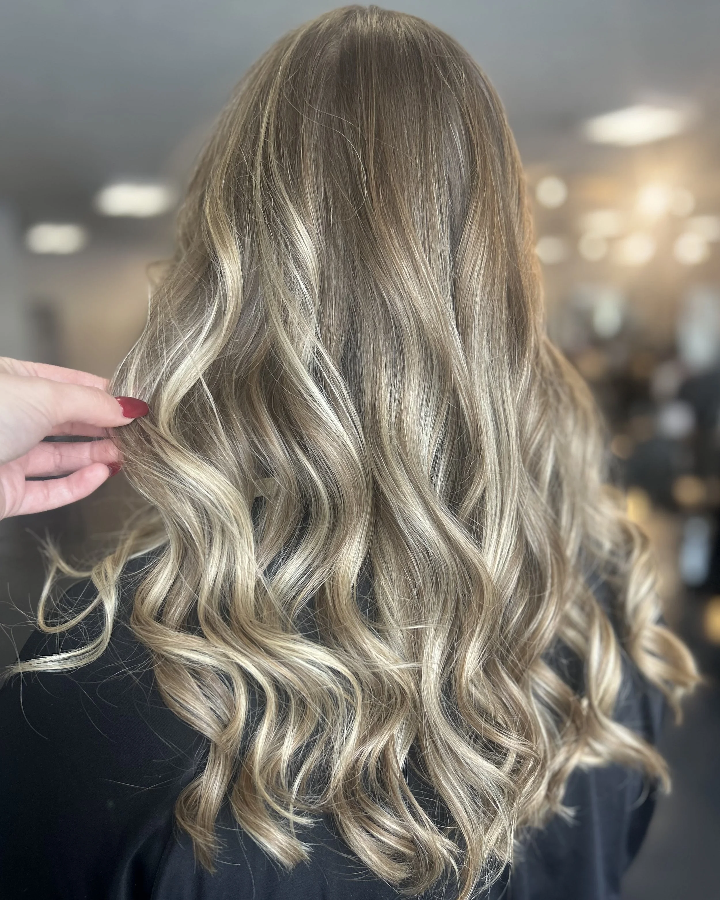 Close-up of a person with long, wavy blonde hair, holding a strand to show the color and texture, against a blurred indoor background.