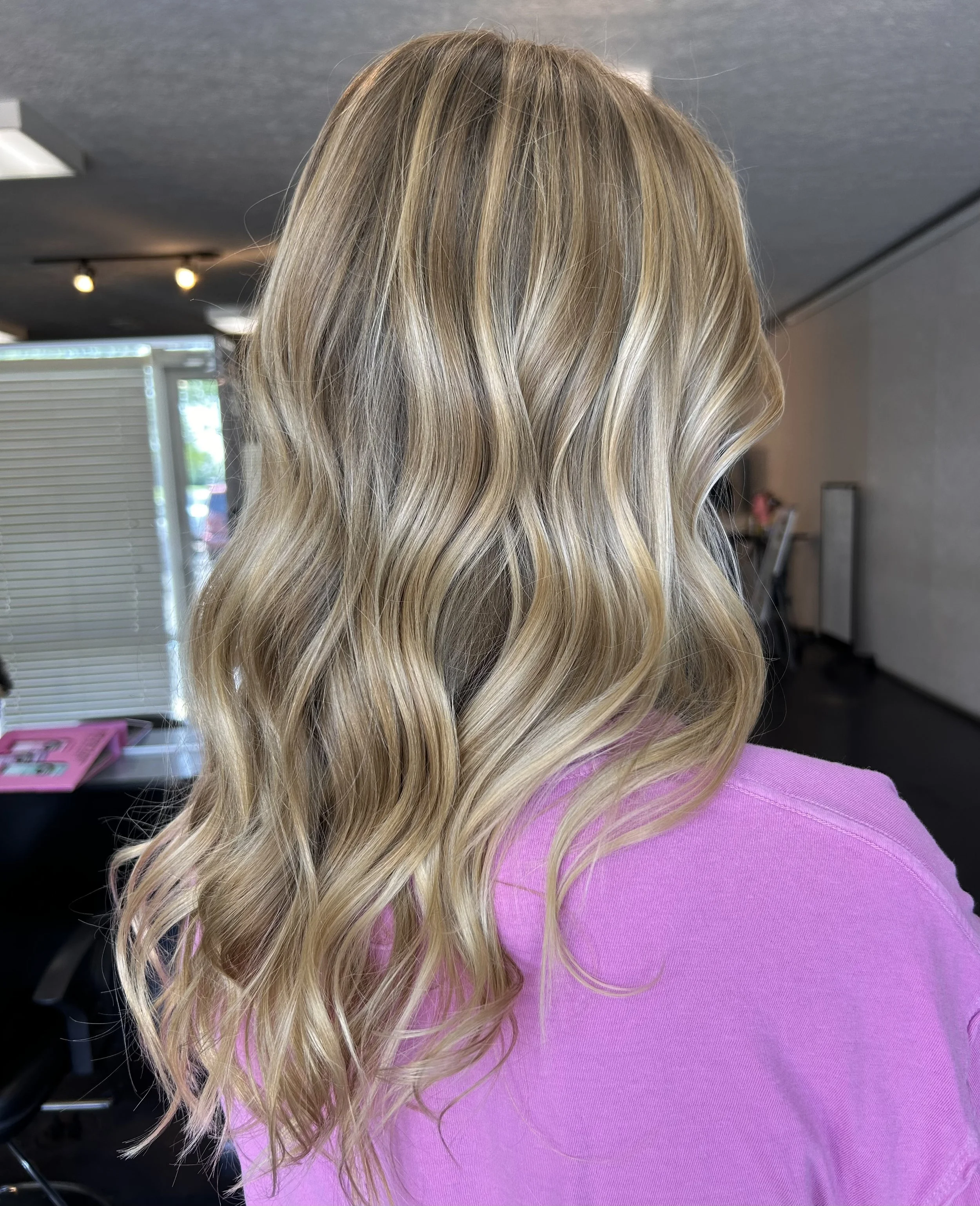 Close-up of a person with long, wavy blonde hair wearing a pink top, standing indoors near a window with natural light and some indoor lighting.