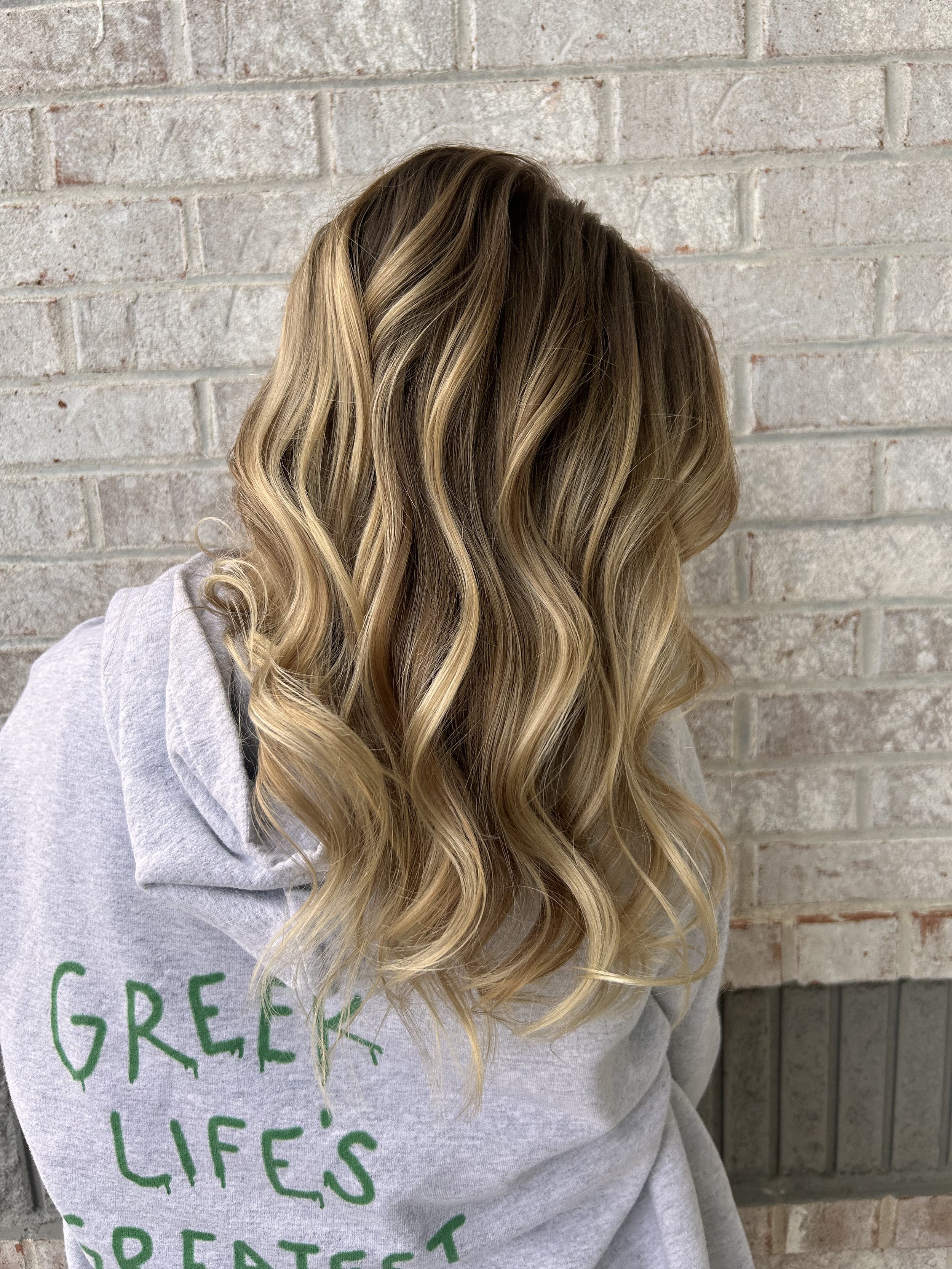 Close-up of a woman with long, wavy blonde hair standing in front of a light-colored brick wall. She is wearing a light gray sweatshirt with green text.