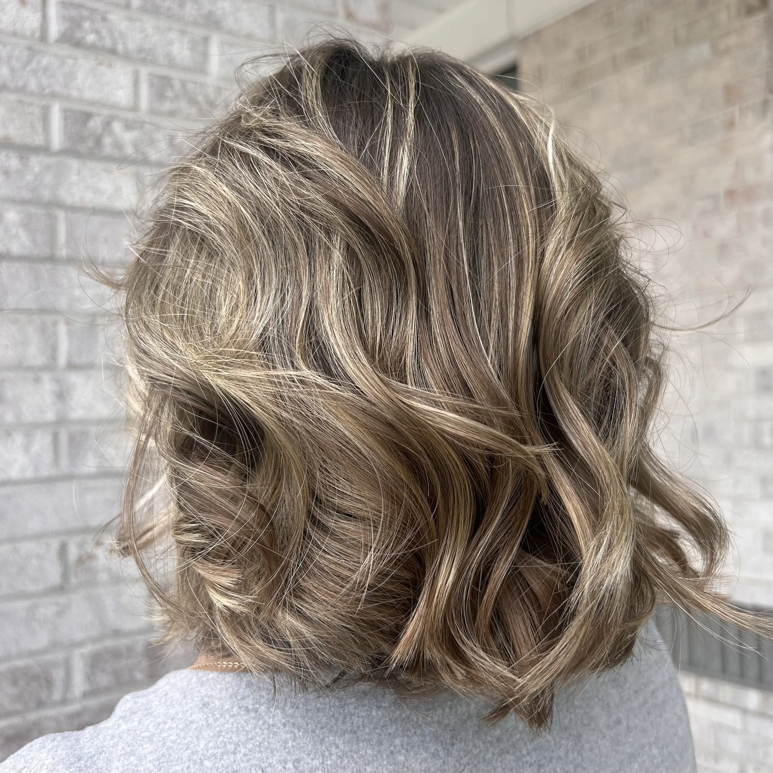 Close-up of a person's hair with light brown and blonde highlights, styled in loose waves, against a gray brick wall background.