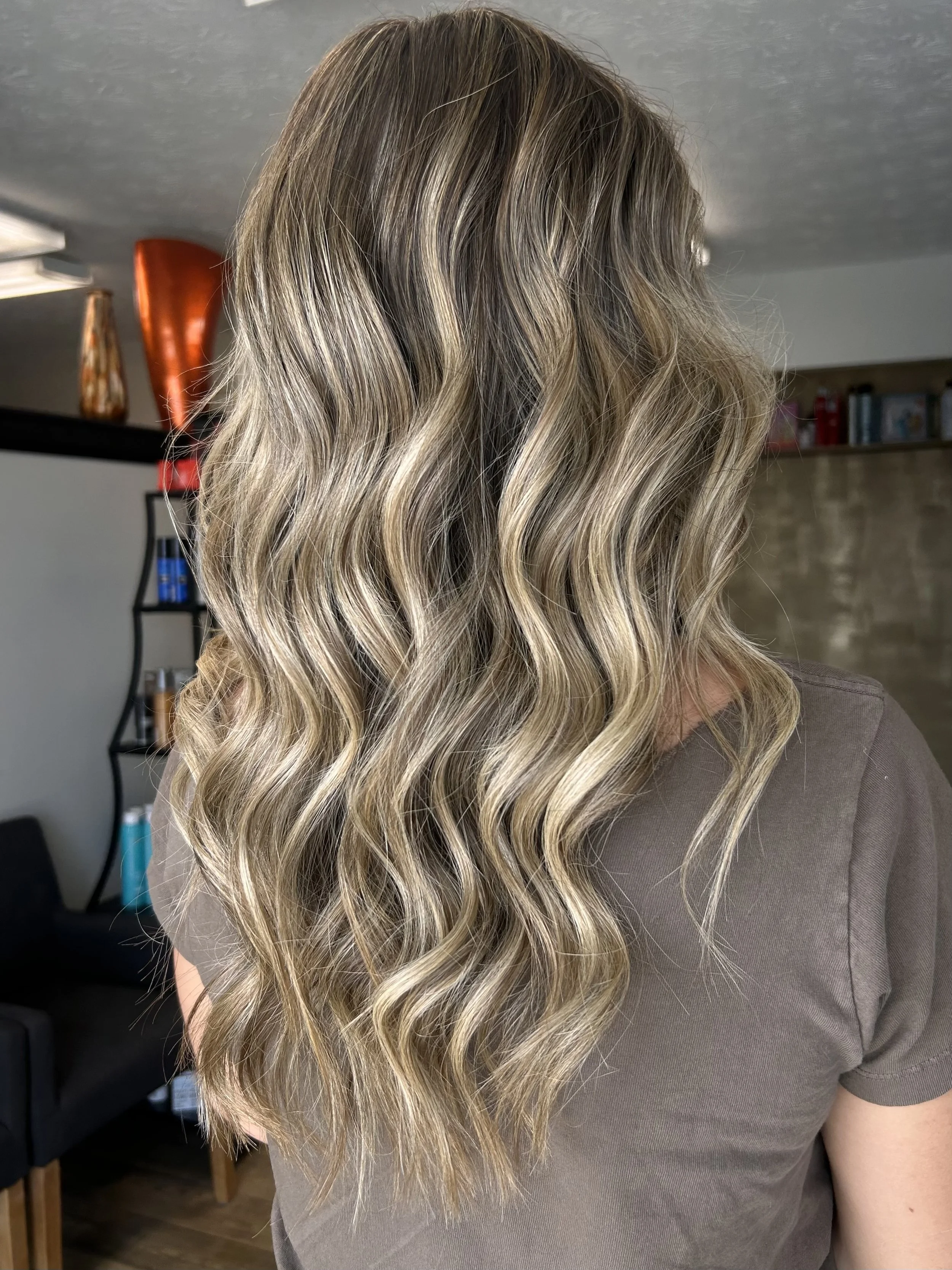 Close-up of someone with long, wavy, blonde hair with highlights, wearing a gray top, indoors with shelves and decorative objects in the background.