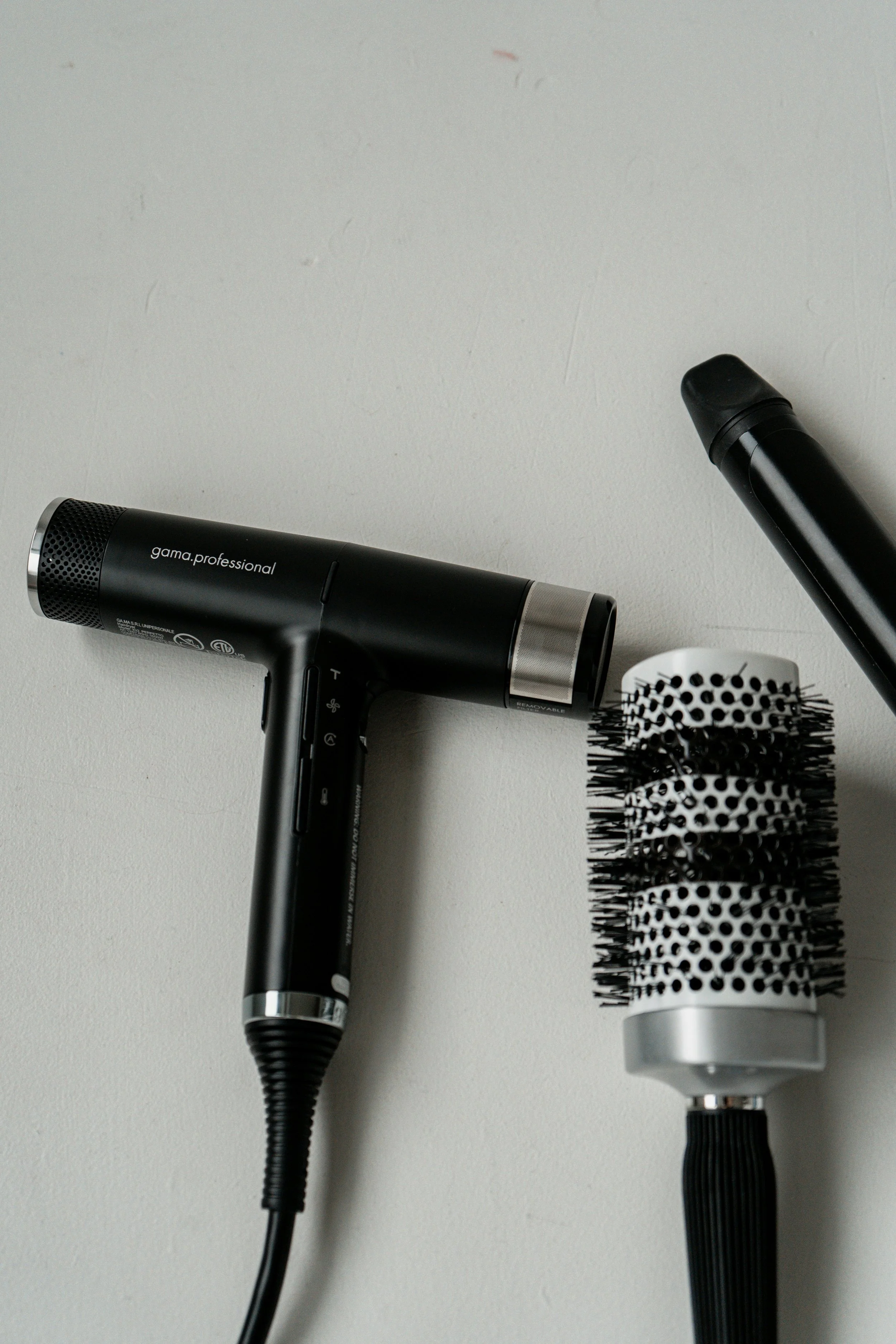 A black hair dryer, a round brush with black bristles and dotted white and black pattern, and a flat hair iron on a light-colored surface.