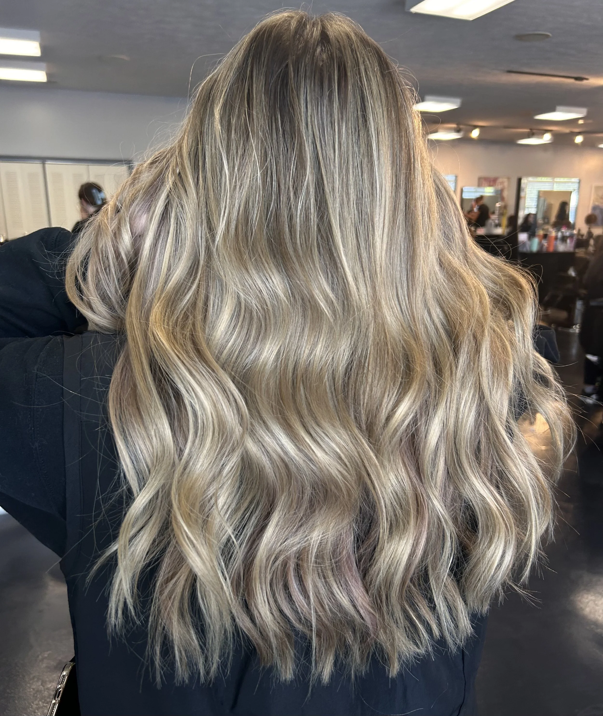 Close-up of a woman with long, wavy blonde hair in a salon, with salon chairs and mirrors in the background.