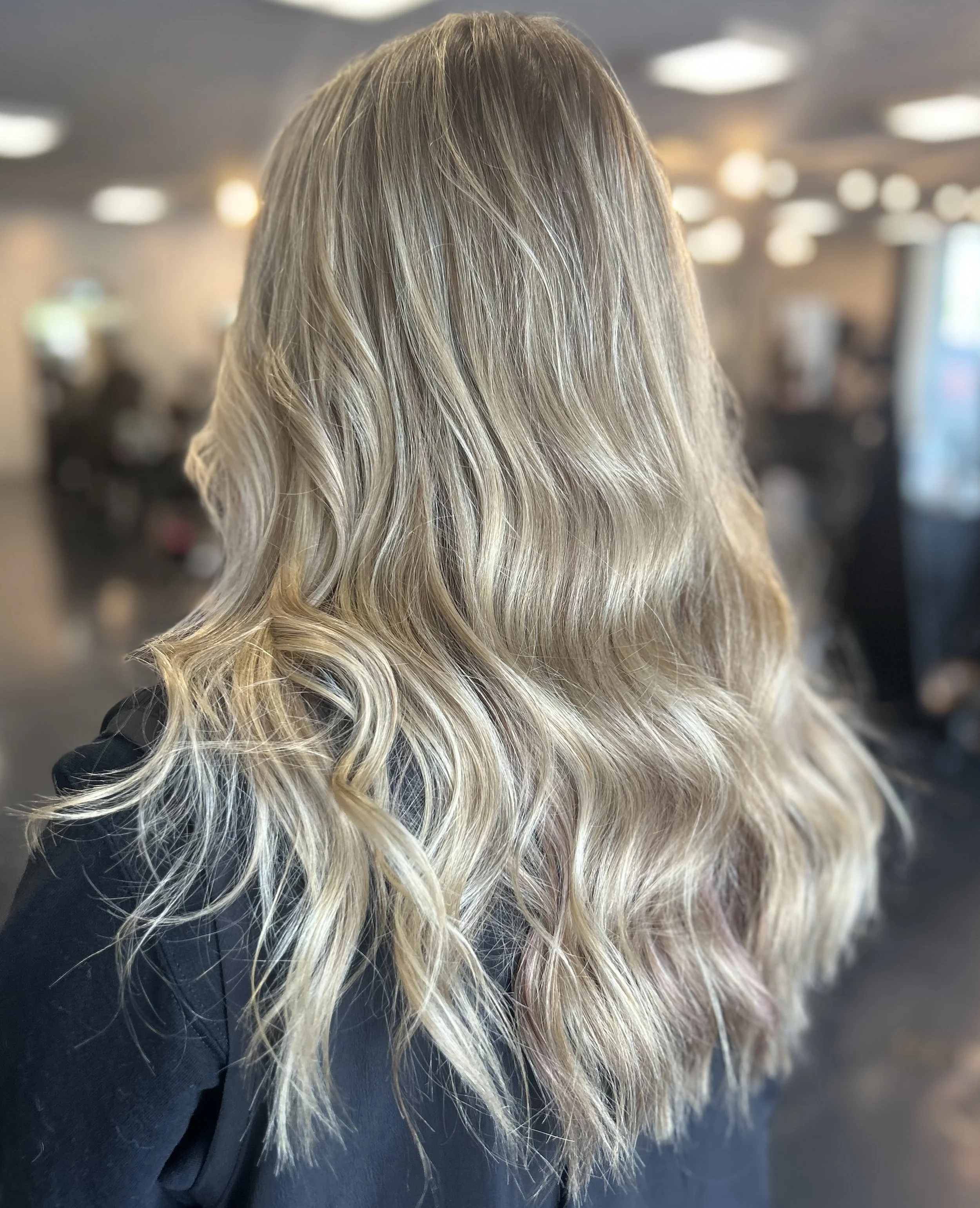 Blonde woman with wavy hair in a salon or barbershop setting, with blurred background of salon interior and lights.