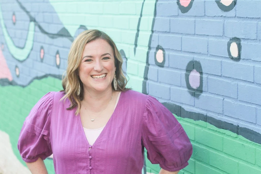 Nashville therapist, Emily Carroll Negi, LMFt, wearing a purple blouse smiling in front of a colorful mural painted on a brick wall.
