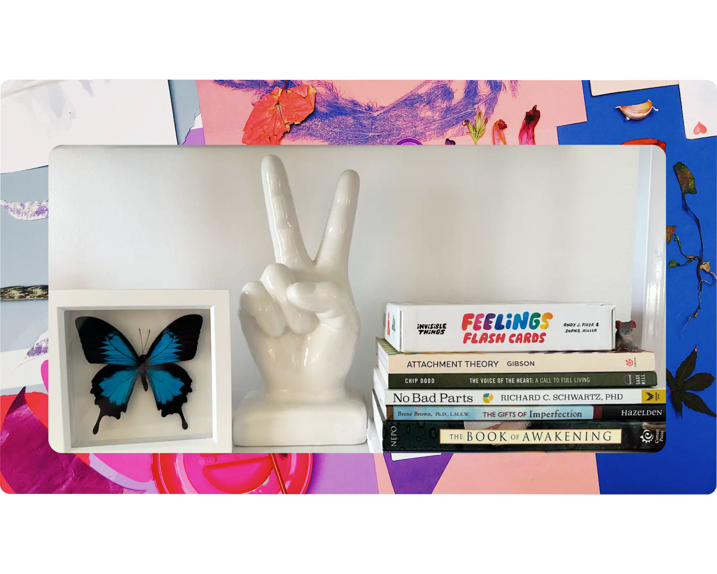 A display shelf with books including 'Feeling Flash Cards', 'Attachment Theory', 'No Bad Parts', and 'The Book of Awakening', a white ceramic hand making a peace sign, and a framed blue butterfly.