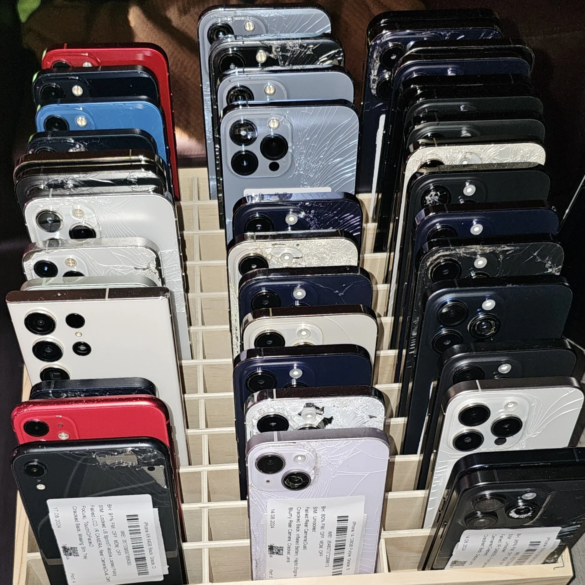 Multiple iPhones and Samsung phones placed on a wooden display stand, some with cracked screens, in various colors including white, black, silver, blue, red, and gold, with barcode tags attached.