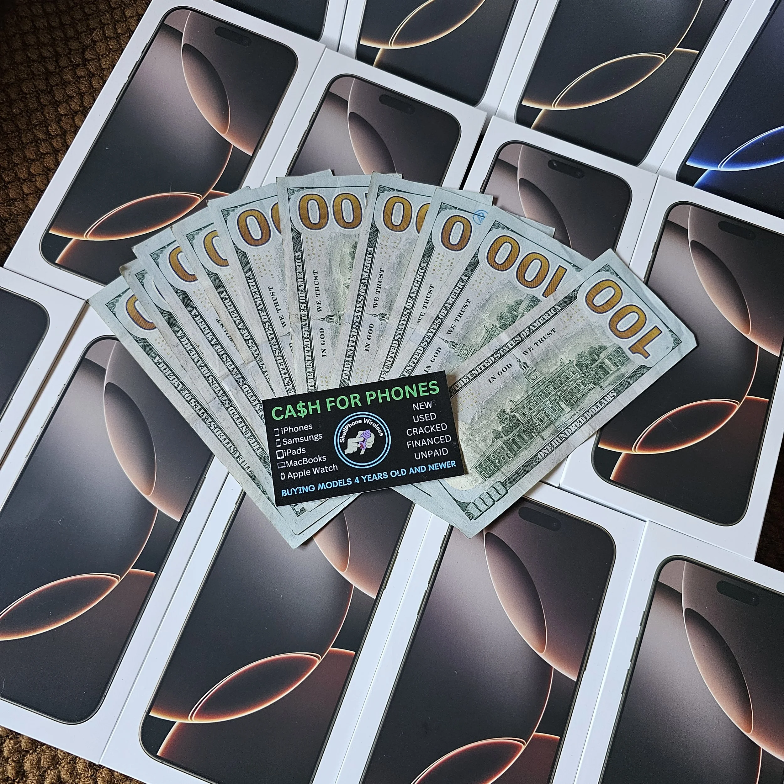 Several sealed boxes of new iPhone 16 Pro Max phones, along with a stack of 100-dollar bills, are spread out on a surface. A flyer advertising cash for phones is also visible.