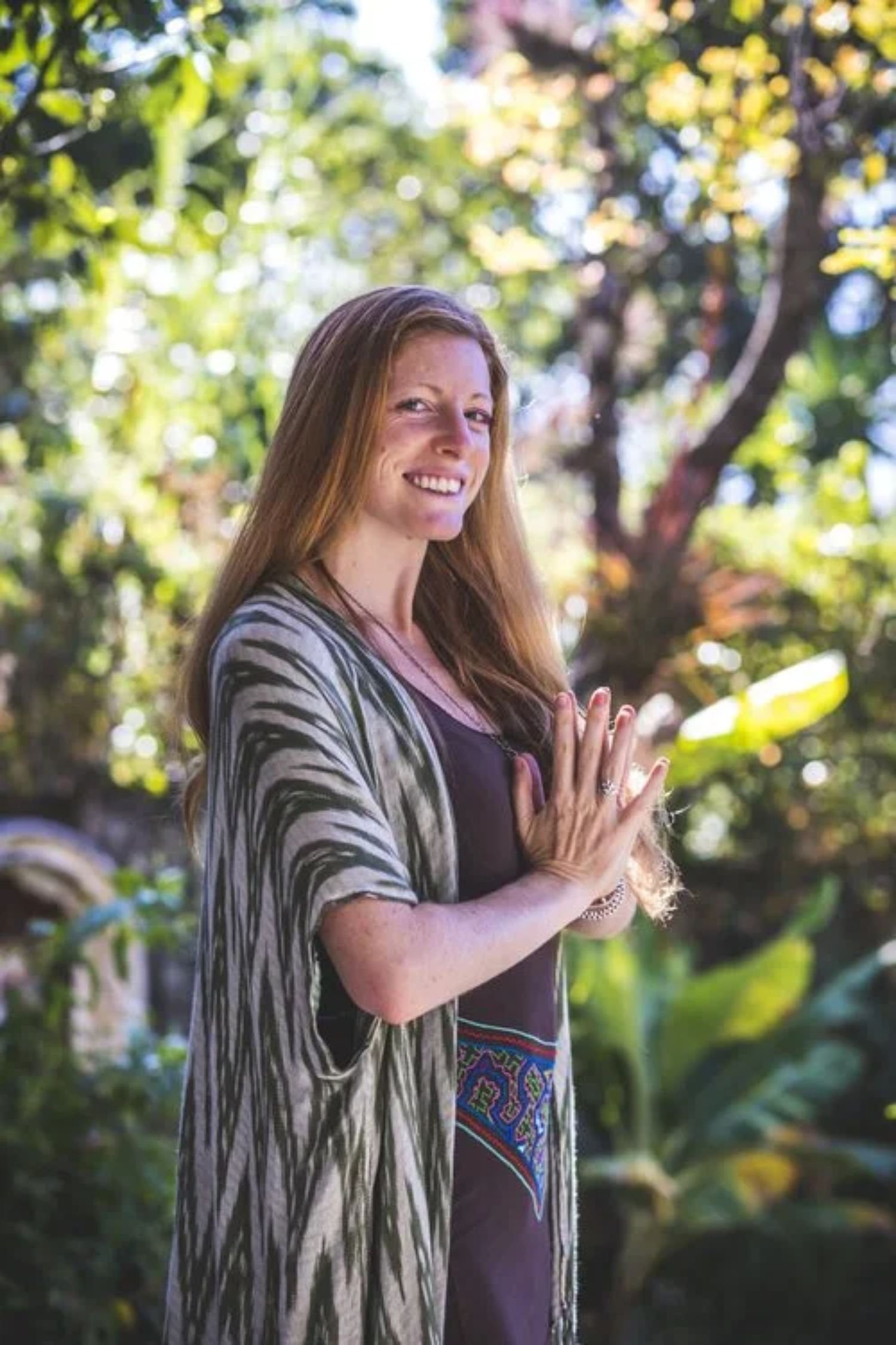 A woman with long reddish hair smiling outdoors, standing with her hands pressed together in a prayer position, in a lush green garden with sunlight filtering through the trees.