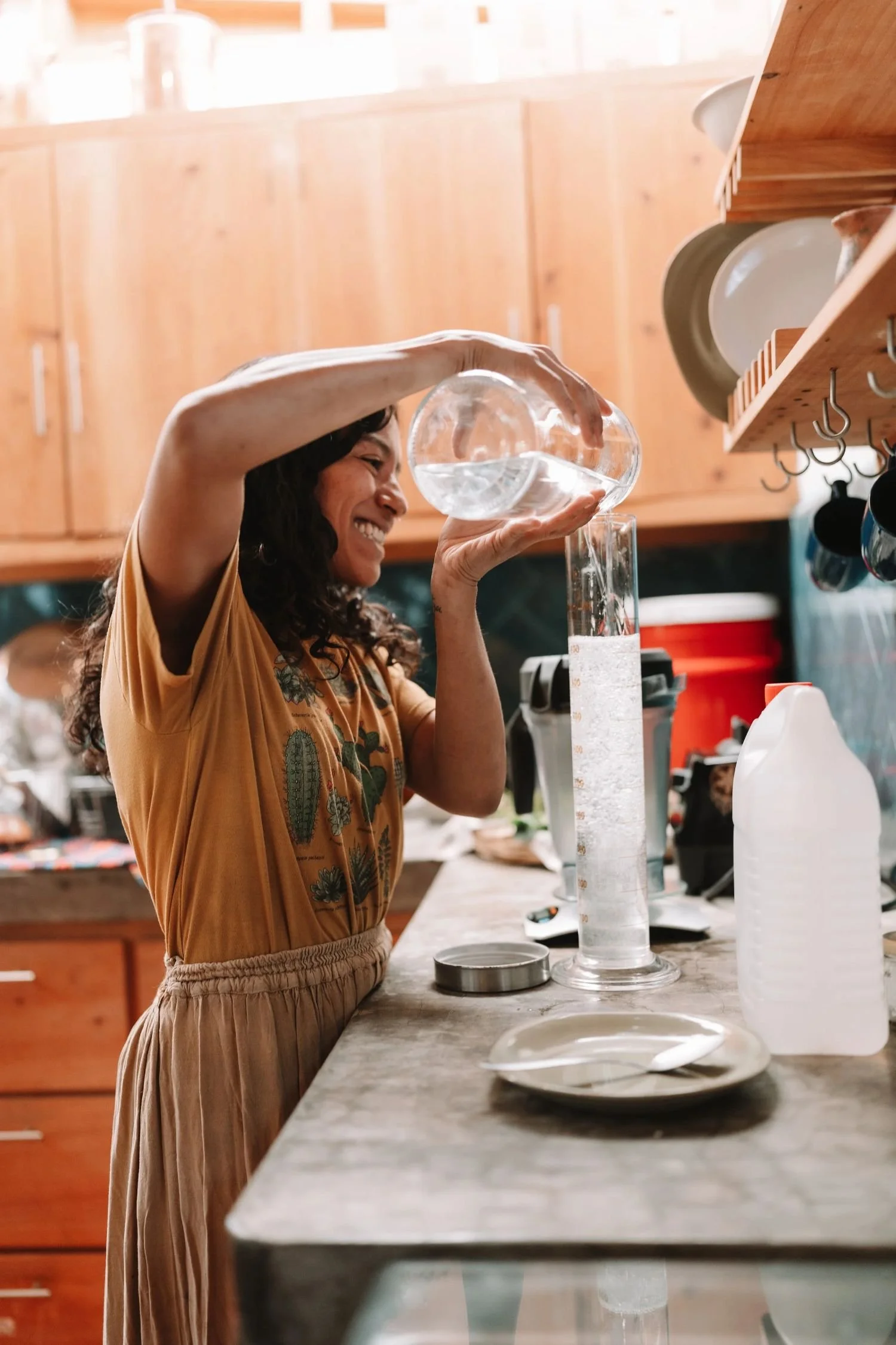 A woman with curly hair smiling and pouring water from a glass pitcher into a test tube in a kitchen with wooden cabinets.