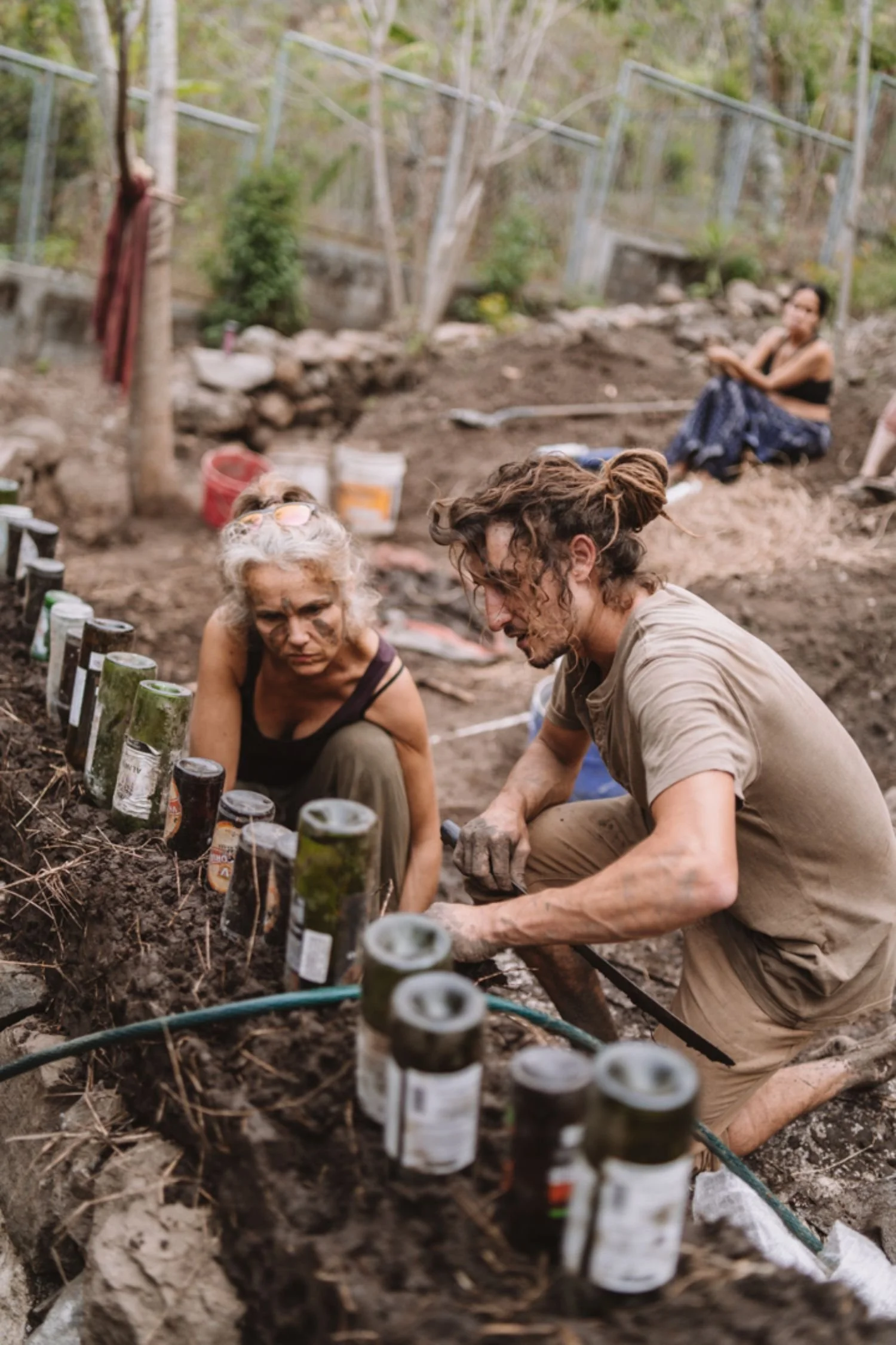 Two people planting seeds in a garden bed surrounded by bottles and gardening tools, with another person sitting in the background among trees.
