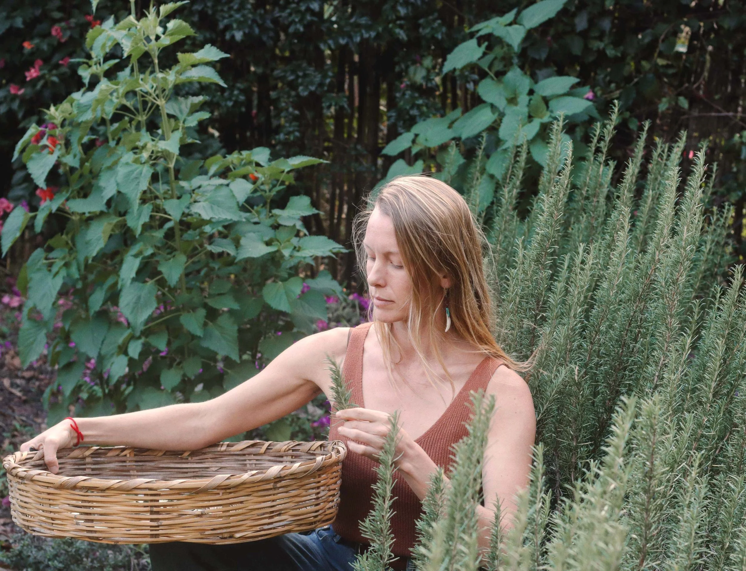 A woman with long, brown hair, wearing a sleeveless brown top, is sitting outdoors in a lush garden with large green leaves, pink and purple flowers, and tall green plants. She holds a woven basket in her left hand, and her eyes are closed, with a peaceful expression.