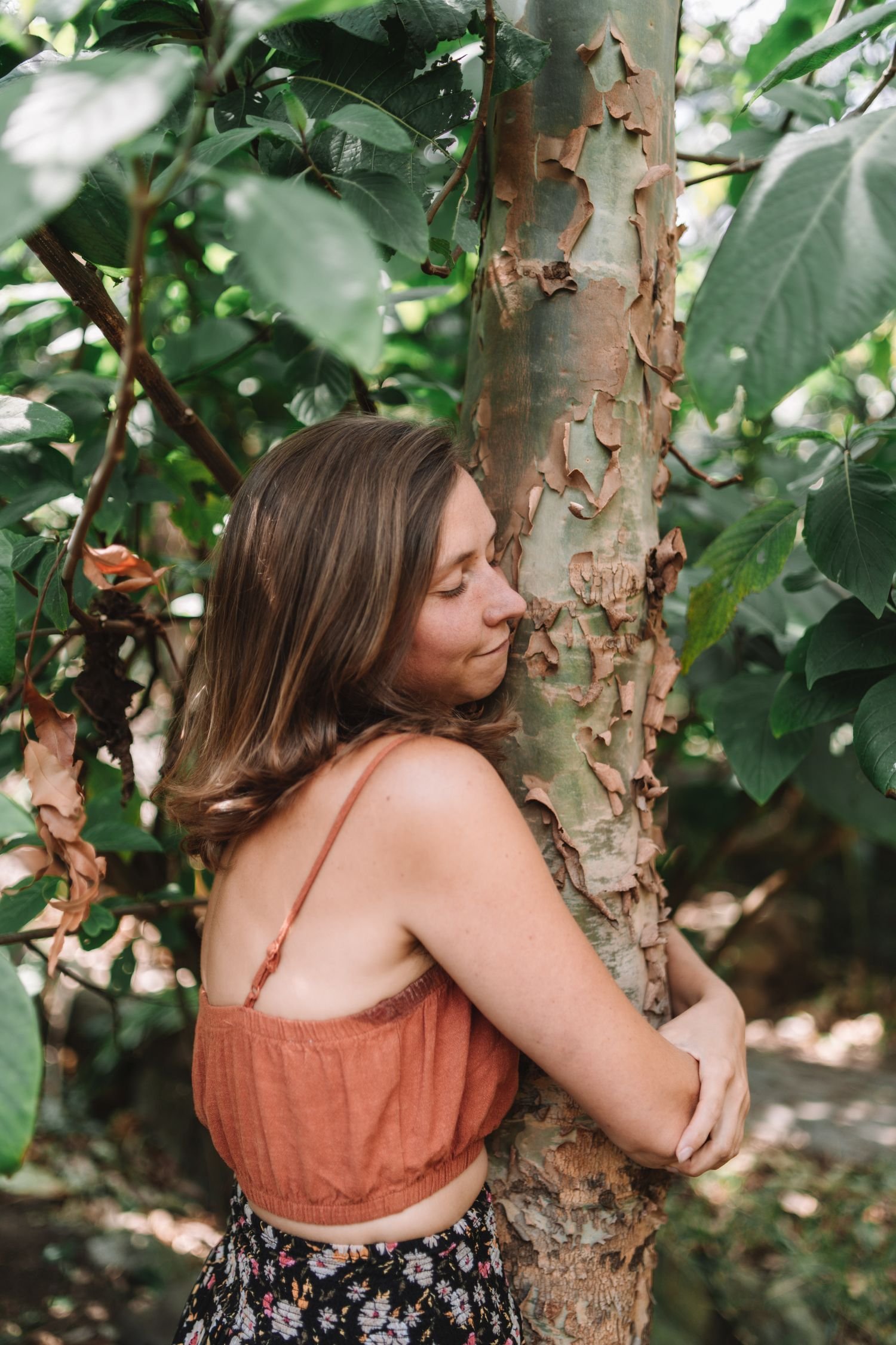 A woman with brown hair in a loose wave, wearing an orange sleeveless top and a black floral skirt, hugging a tree in a dense green forest, with her eyes closed and a peaceful expression.