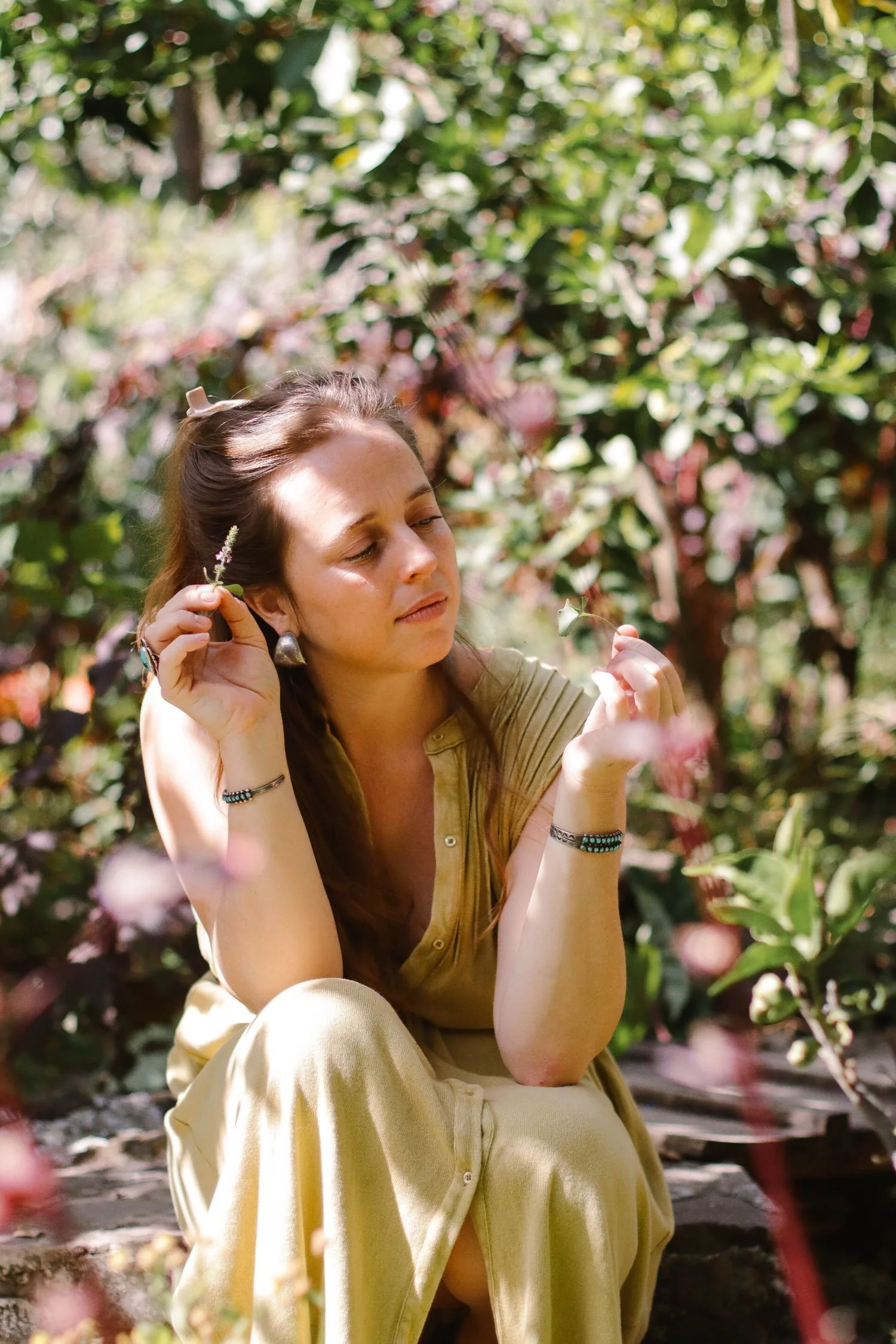 A woman with long brown hair sitting outdoors among greenery, holding a small flower near her face with her eyes closed.