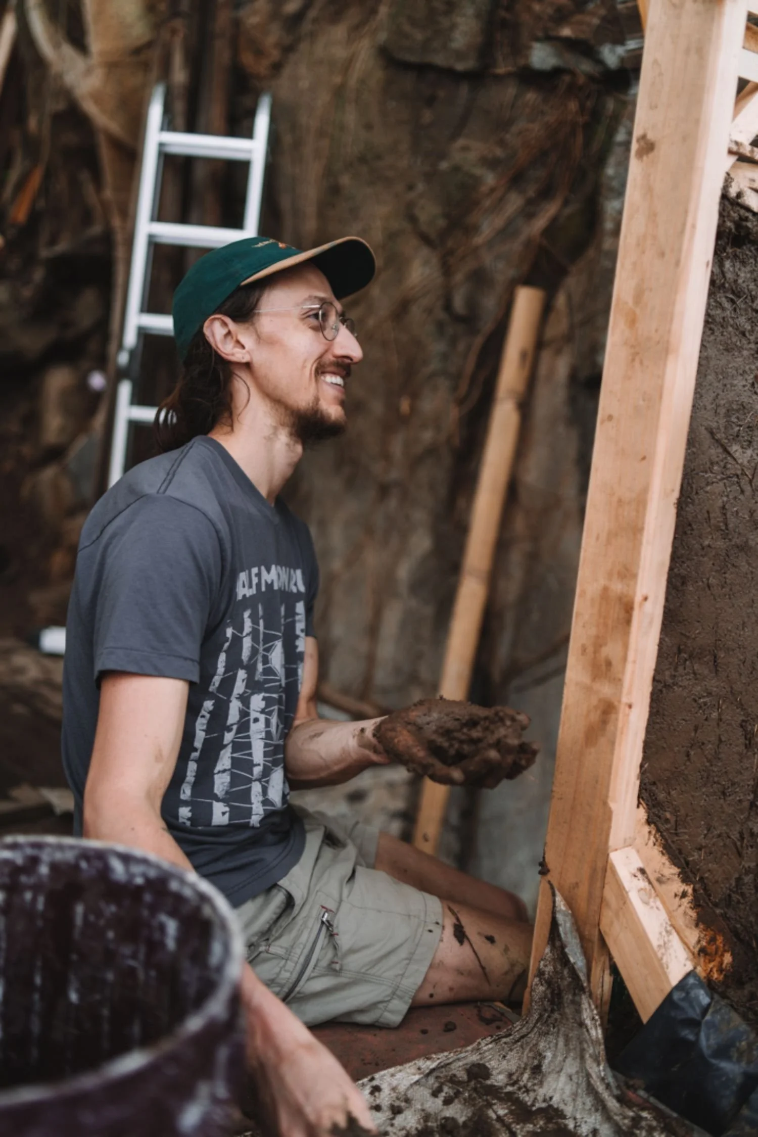 A man with glasses, long hair, and a beard is working in a construction or workshop setting. He is wearing a green cap, a gray T-shirt, and khaki shorts. The man is holding a clump of soil or clay and is smiling. Behind him, there is a wooden structure, a ladder, and a rough rock or earth wall.