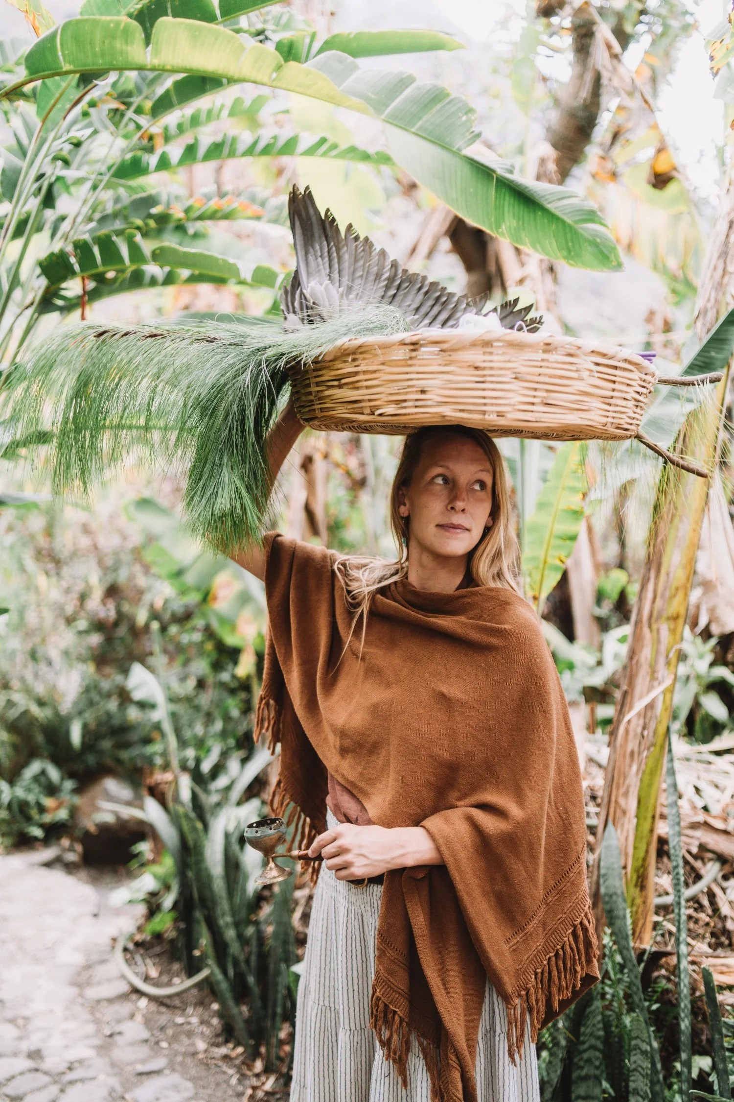 A woman outdoors in a lush, green garden, holding a basket made of woven wicker on her head, surrounded by large leafy plants.