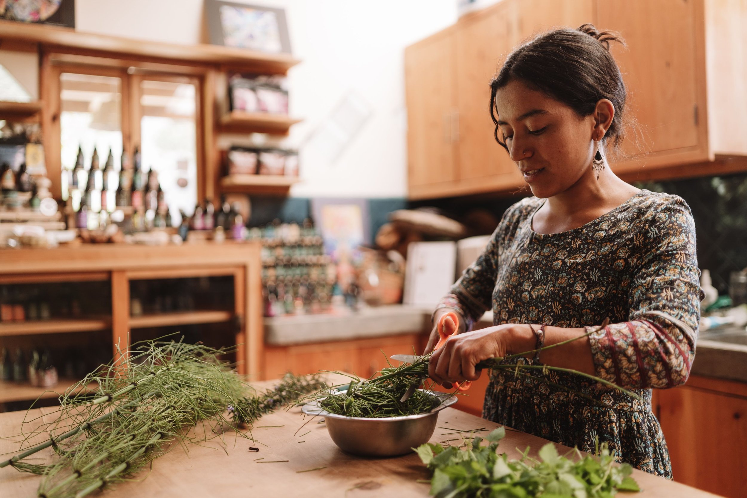 Woman in floral dress preparing herbs or plants in a rustic kitchen with wooden shelves and cabinets.