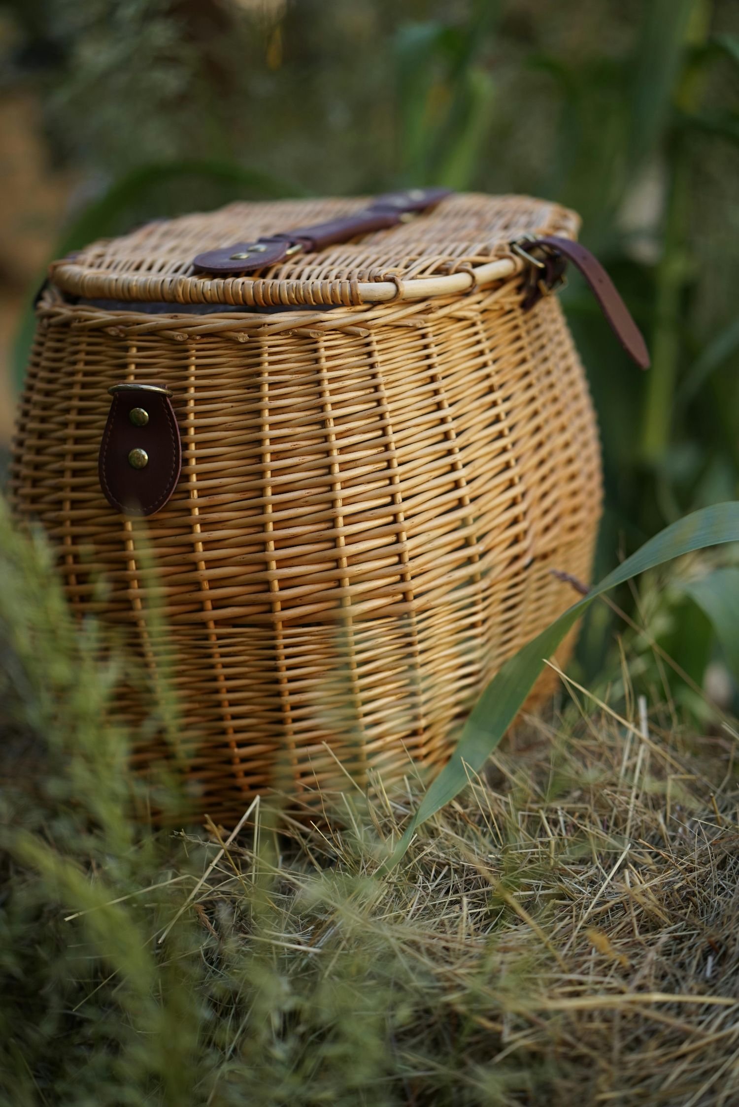 A wicker bicycle basket with leather straps on grass and plant background.