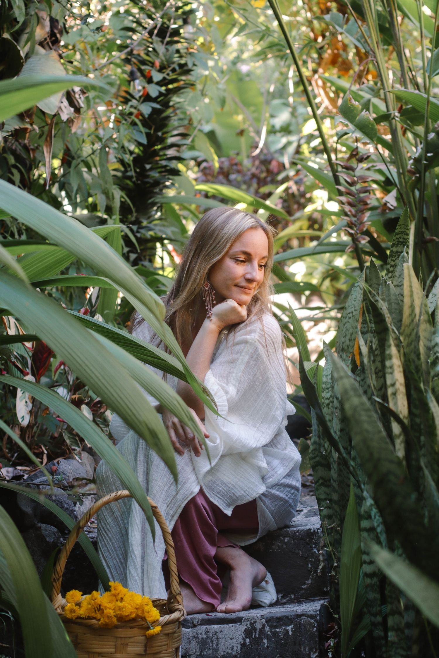 A woman crouching on stone steps among lush green plants, holding her chin with her hand and smiling softly. A basket of yellow marigold flowers sits nearby.