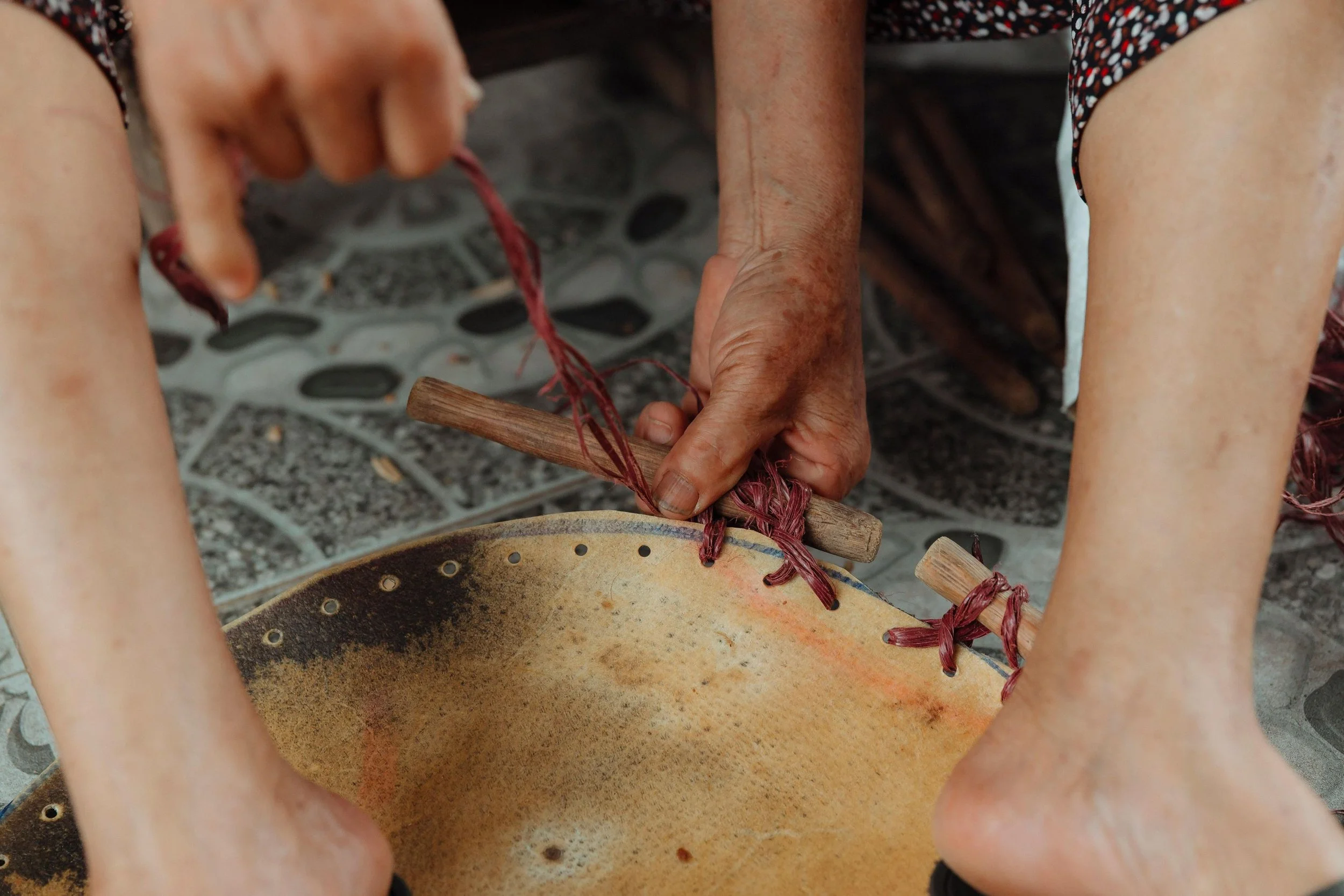 Close-up of a person playing a traditional drum with red strings attached, with their hands holding sticks. The person is wearing a patterned shirt and shorts, and the drum is on a tiled surface.