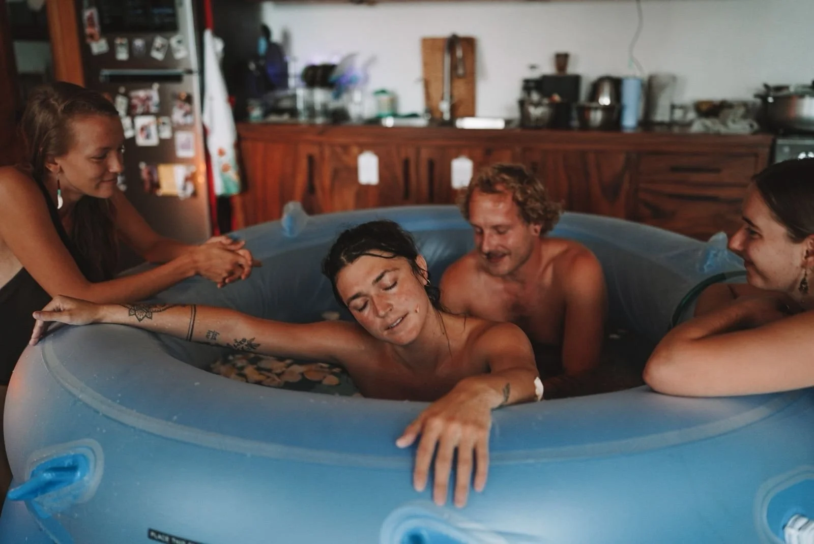 People celebrating a person's baptism in a blue inflatable pool indoors, with the person in the center being baptized and others smiling around.