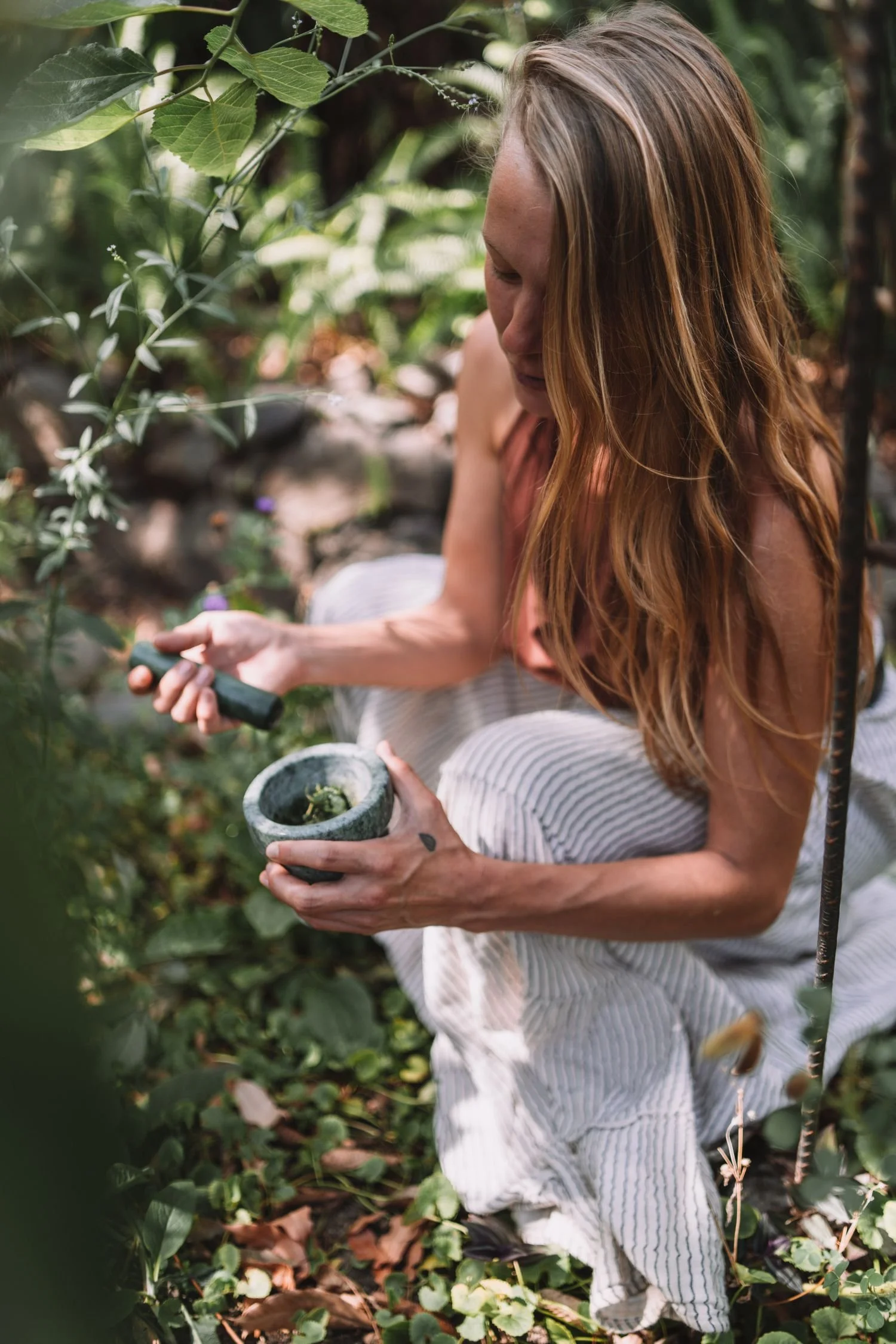 Woman with long reddish hair sitting amidst green plants, holding a stone mortar and pestle, in garden or natural outdoor setting.