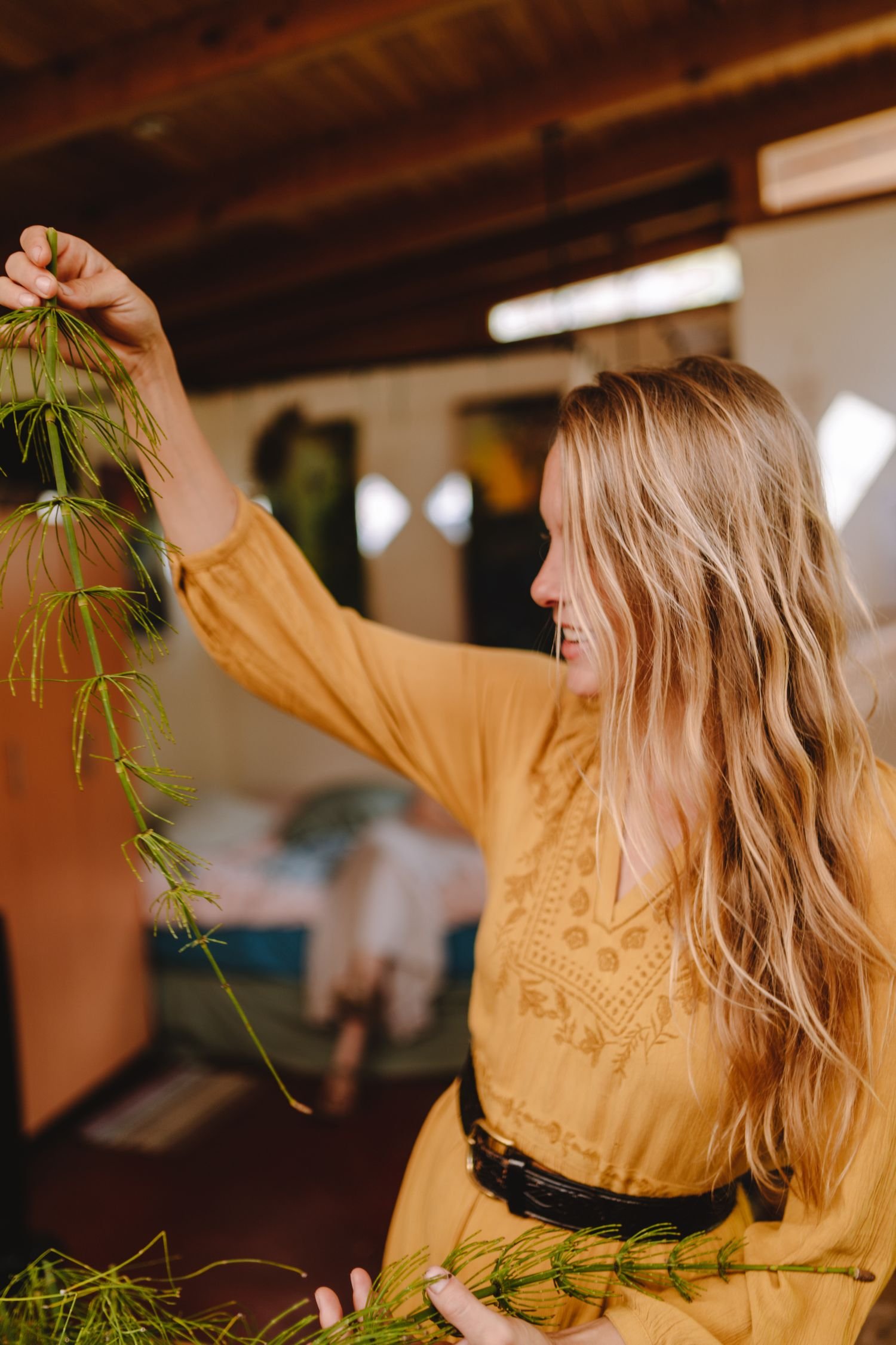 A woman with long wavy blonde hair wearing a yellow embroidered blouse is arranging a green leafy plant indoors.
