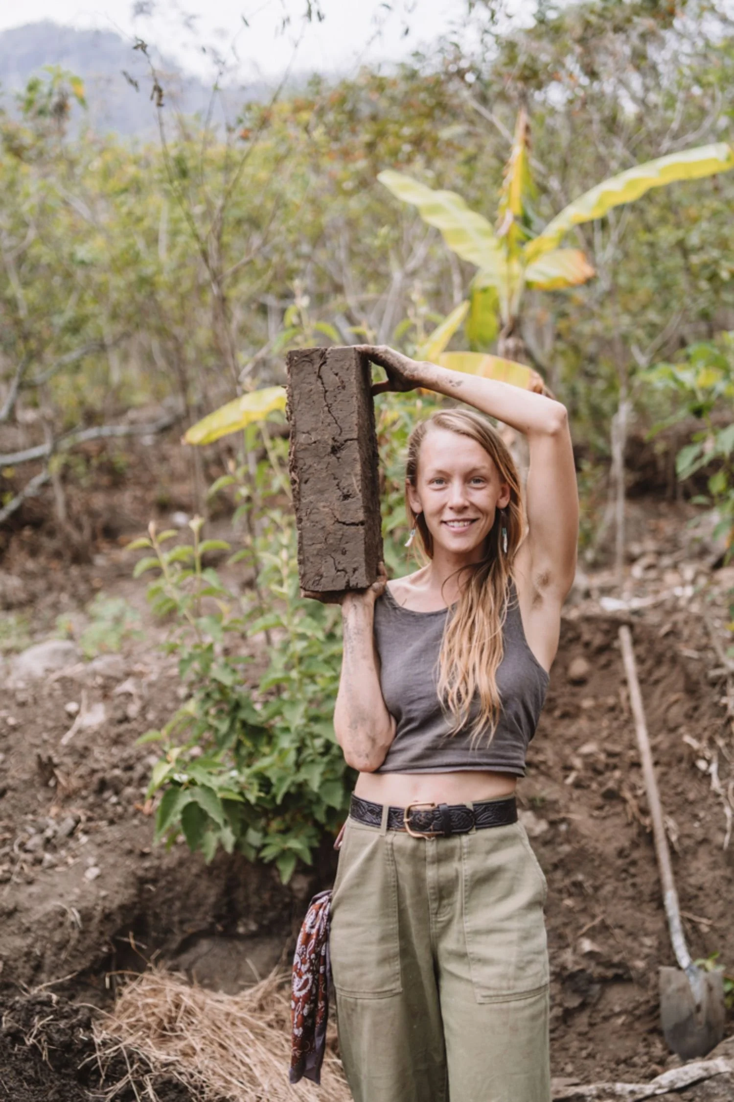 A woman with long, wavy hair, wearing a sleeveless top and khaki pants, is standing outdoors in a natural setting with plants and trees. She is smiling and holding a piece of wood or log over her shoulder.