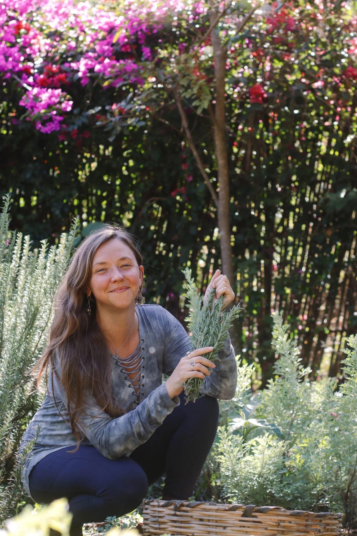 Young woman with long brown hair, smiling, holding sprigs of lavender in a garden with green plants and pink and red flowering bushes in the background.