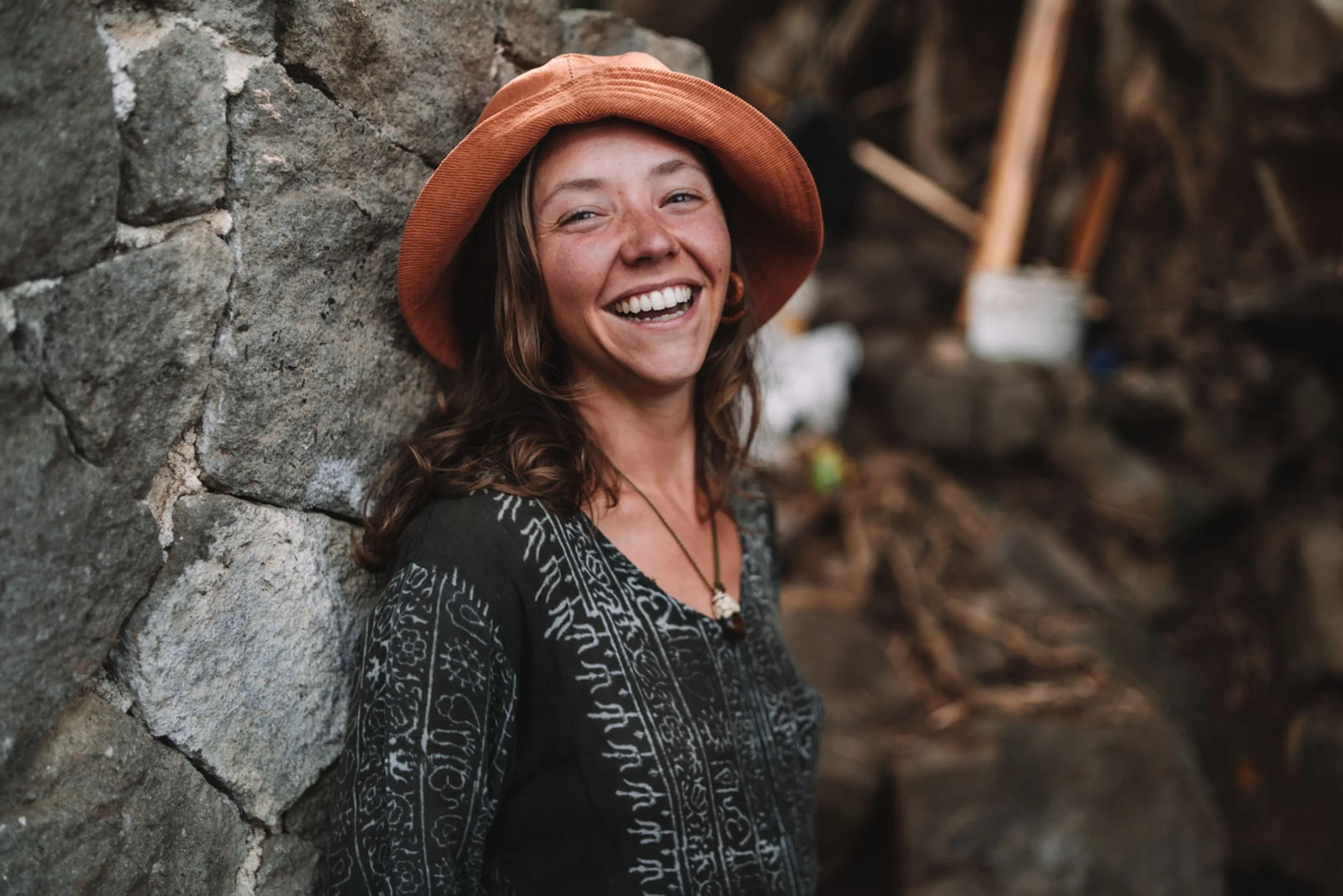 A smiling woman with long brown hair wearing a tan wide-brimmed hat and a black dress with white intricate patterns, standing against a stone wall inside a rustic wooden structure.