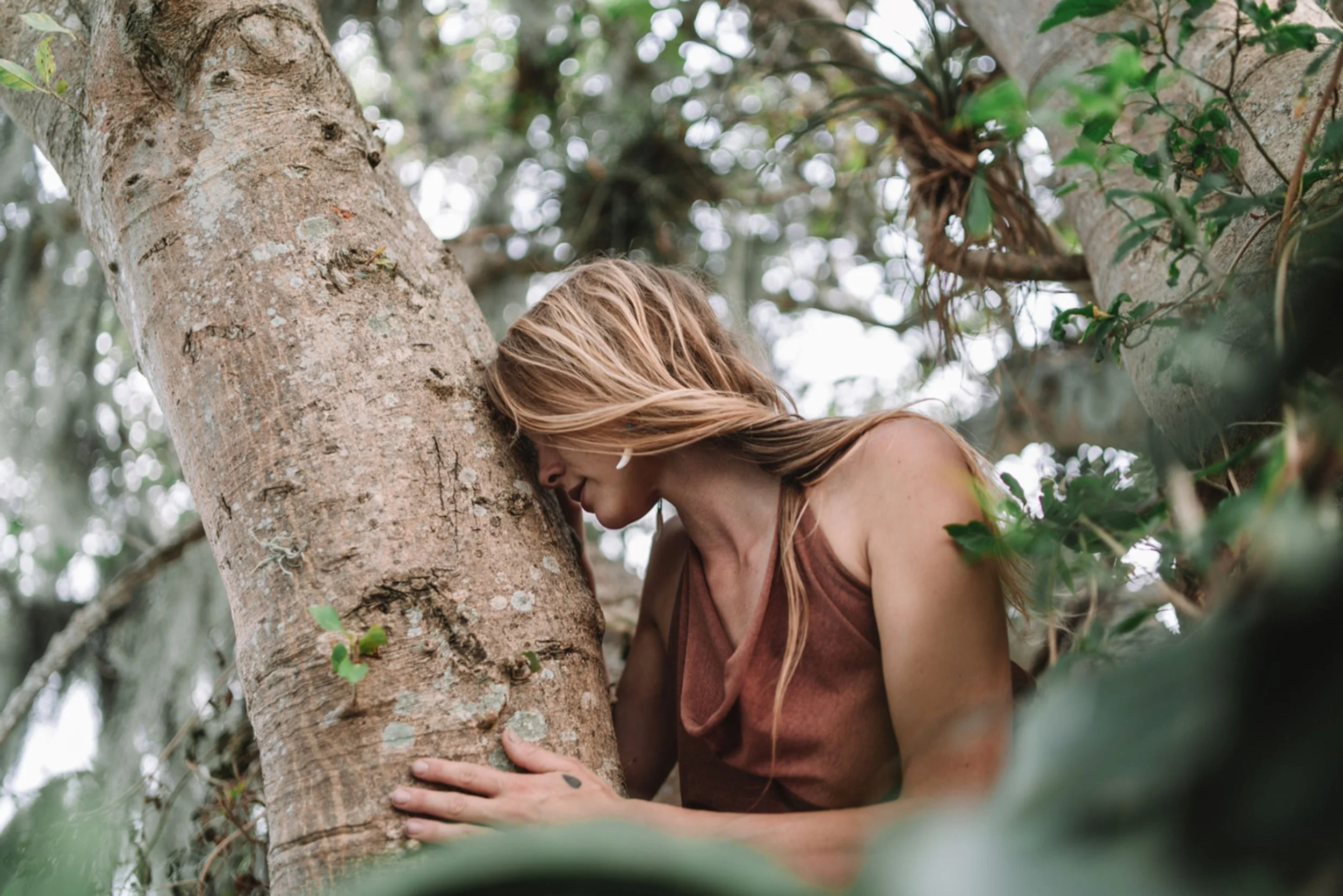 A woman with long blonde hair leaning against a tree trunk in a lush green forest, surrounded by foliage.