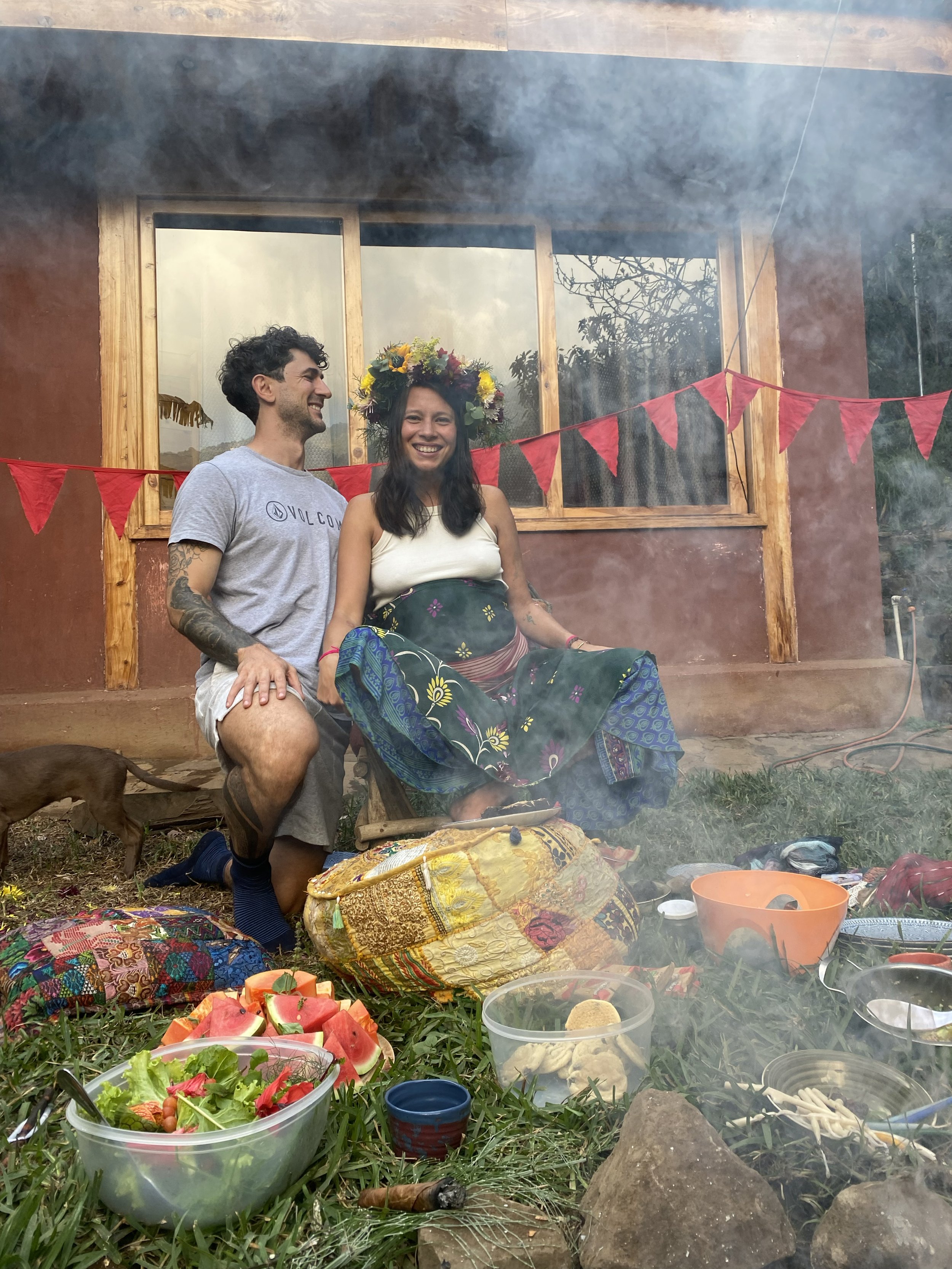 A woman and a man smiling at each other during a celebration outside a rustic house with red bunting. The woman is wearing a floral crown and sitting in a rocking chair. The area is decorated with colorful textiles, food, and drinks, with a fire nearby.