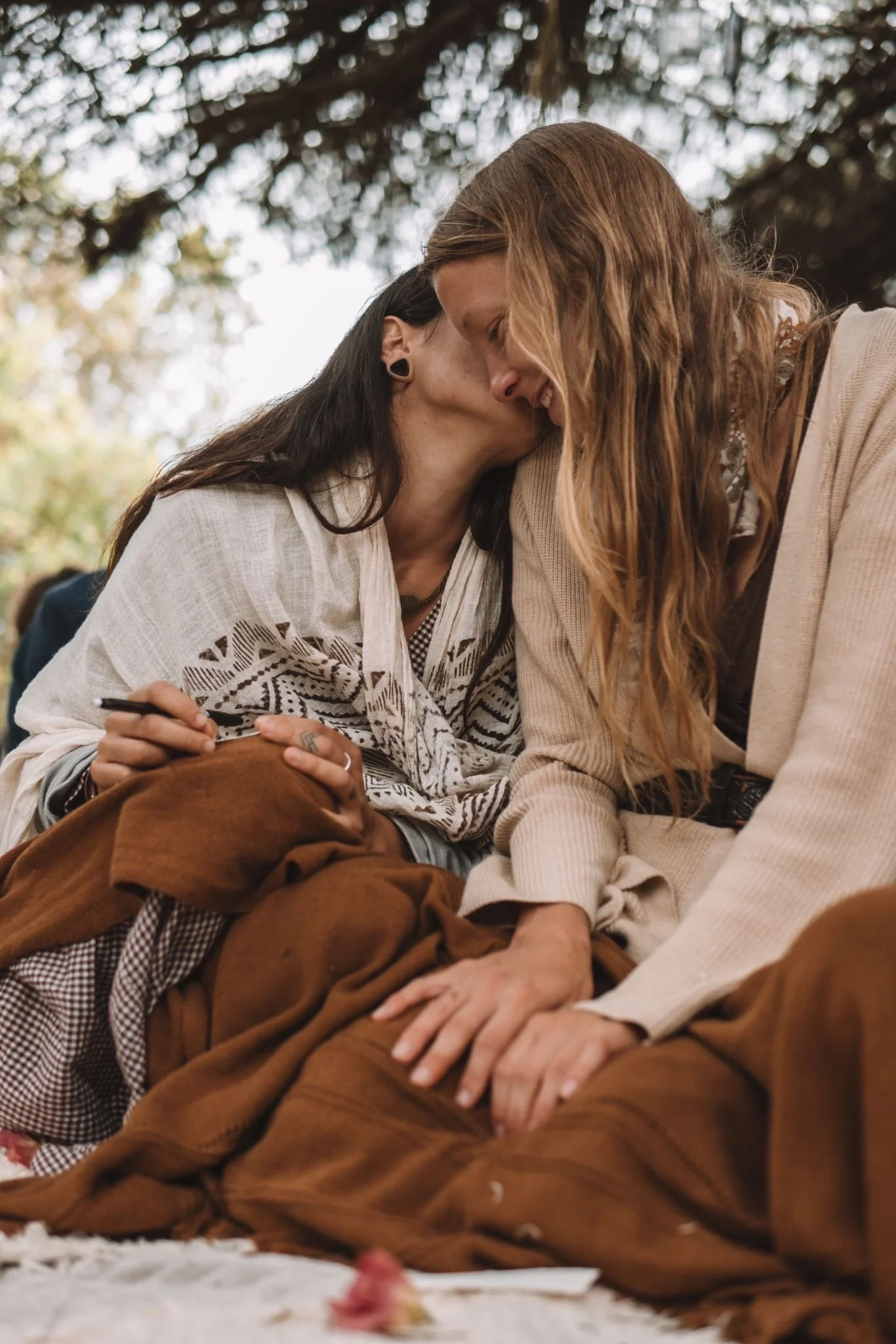 Two women sharing an intimate moment, forehead to forehead, outdoors under a tree, with one woman holding a pen and paper, dressed casually.