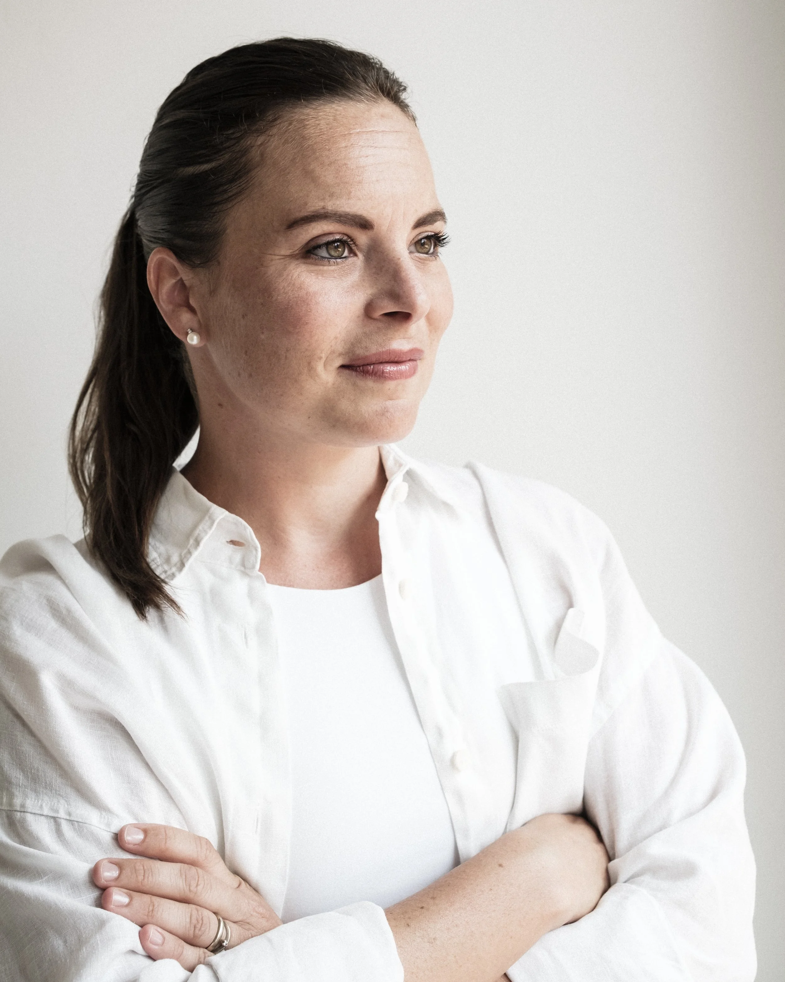 Ritratto di una donna con capelli castani lunghi e occhi verdi, indossa una camicia bianca, sorridente, con le braccia incrociate e un orecchino di perla, sfondo neutro.