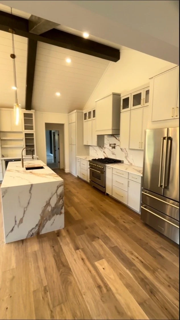 Modern kitchen with white cabinets, marble countertops, and a stainless steel refrigerator. There is a marble island with a sink, hardwood floors, and a vaulted ceiling with dark beams.