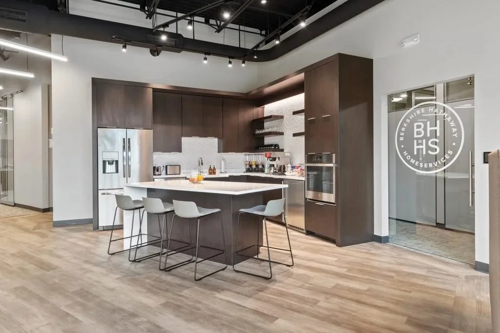 Modern kitchen with dark wooden cabinets, stainless steel appliances, white island counter with four gray bar stools, and light wood flooring.