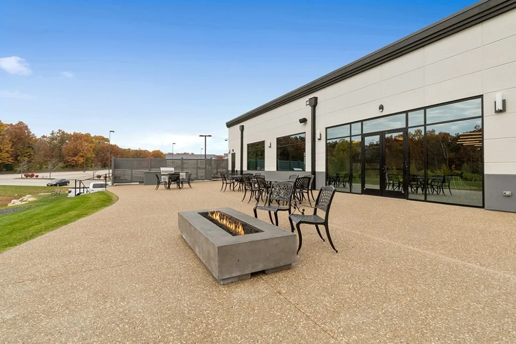 Empty outdoor patio area with tables and chairs, a modern fire pit, adjacent to a building with large windows, with trees and parking lot in the background on a clear day.