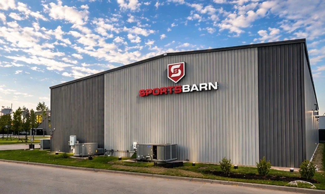 Exterior view of a large metal sports store named Sports Barn with a red and white logo, against a partly cloudy sky.