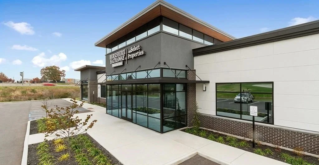 Modern commercial building with glass entrance and signage for Berkshire Hathaway Select Properties, with parking lot and landscaped area in front.