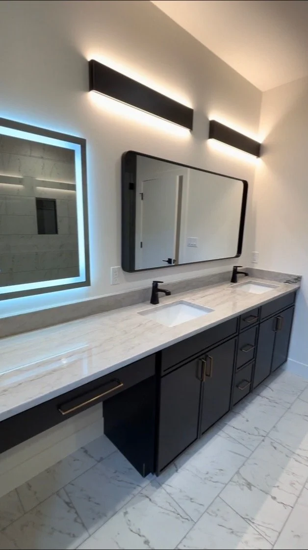 Modern bathroom vanity with a marble countertop, two sinks, black cabinets, and a large mirror with black trim. There are two horizontal black wall lights above the mirror, and a backlit mirror with a blue glow on the side.