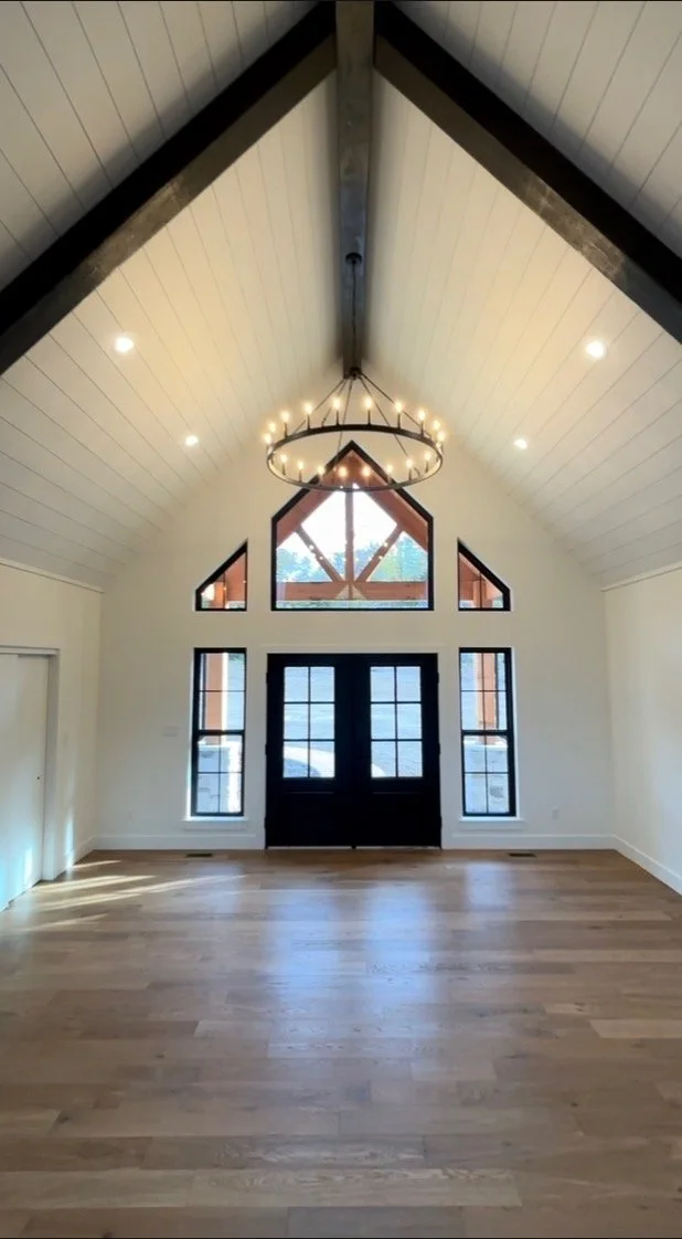 Empty room with vaulted ceiling, large windows, black double doors, and a circular chandelier.