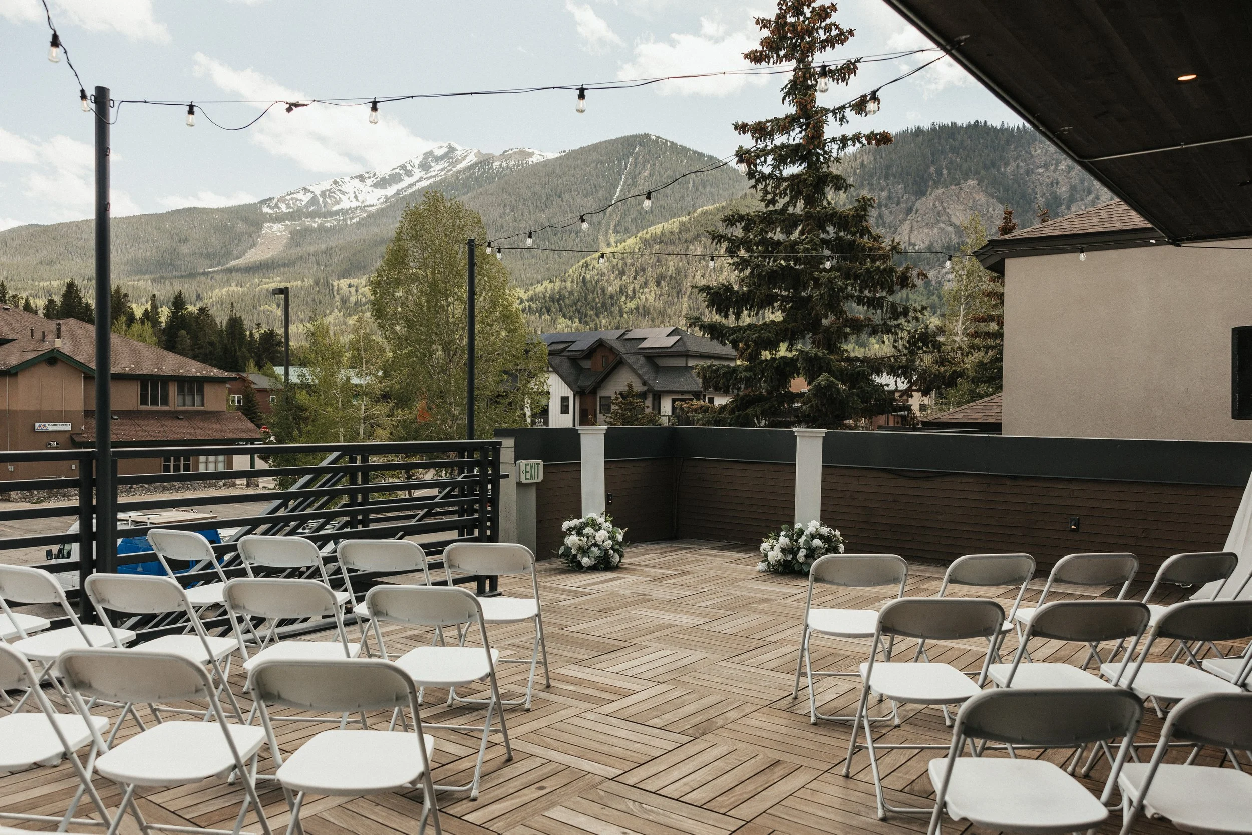 Outdoor venue with rows of white chairs on a wooden deck, decorated with flower arrangements, overlooking a scenic mountain landscape and trees, under a partly cloudy sky.