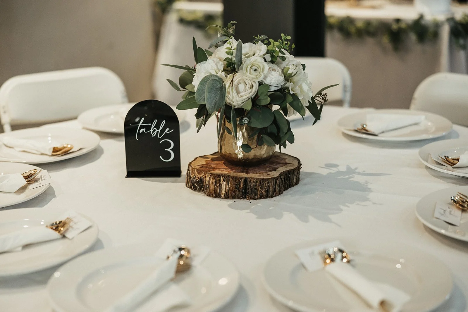 Elegant wedding table setting with white plates, gold cutlery, a flower centerpiece in a gold vase on a tree trunk slice, and a black table number sign reading 'Table 3'.