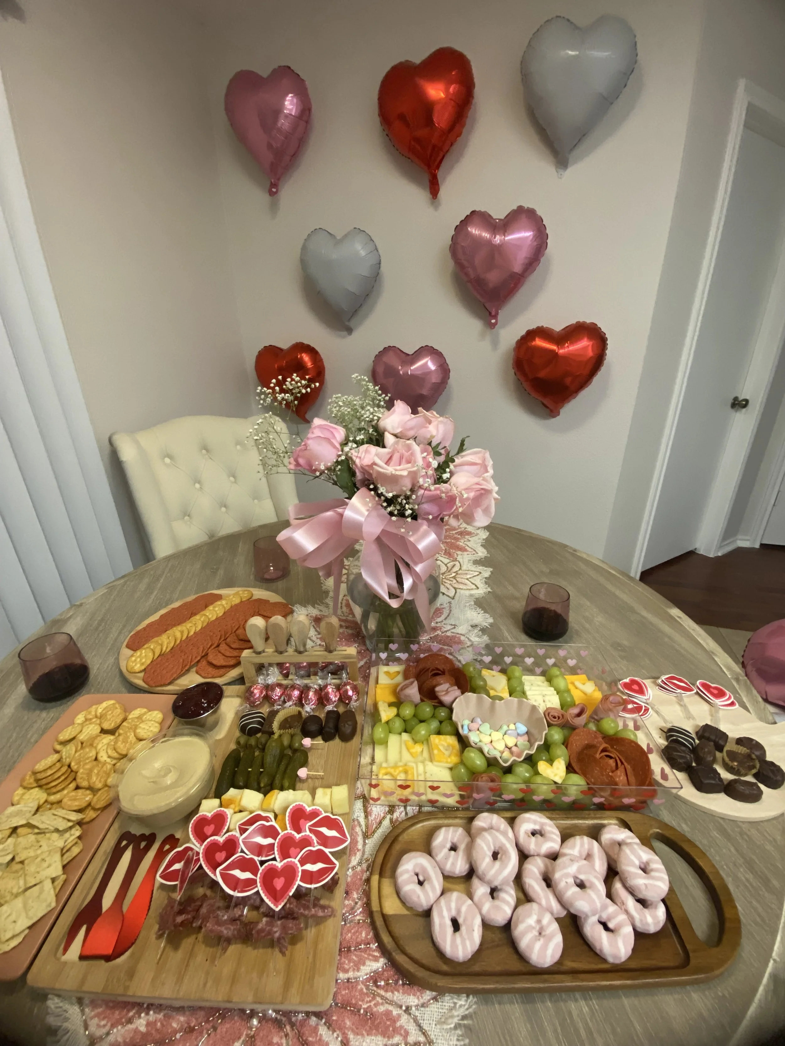 Valentine's Day themed table with heart-shaped balloons in pink, red, and silver, and a bouquet of pink roses. The table has various treats including donuts, cookies, chocolates, cheese, grapes, and decorative heart-shaped picks.