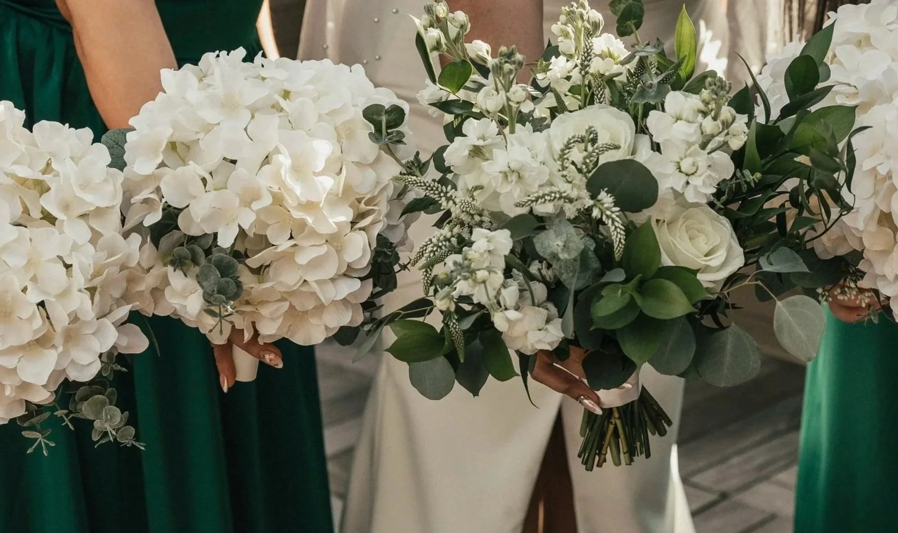 A person holding a bouquet of white flowers including roses and hydrangeas, with green leaves and small white accents.