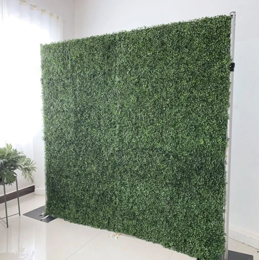 Green artificial hedge panel standing indoors near a potted plant and a window with sheer curtains.