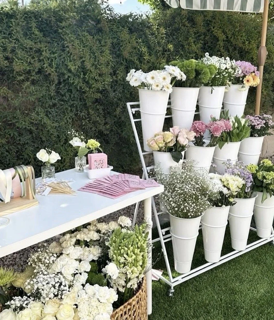 A display of white and pastel-colored flowers arranged in white flower pots, with some green foliage. There is a white table with pink pamphlets, small vases, and decorative items next to the flower display, set outdoors with green hedge background and a partial striped umbrella.