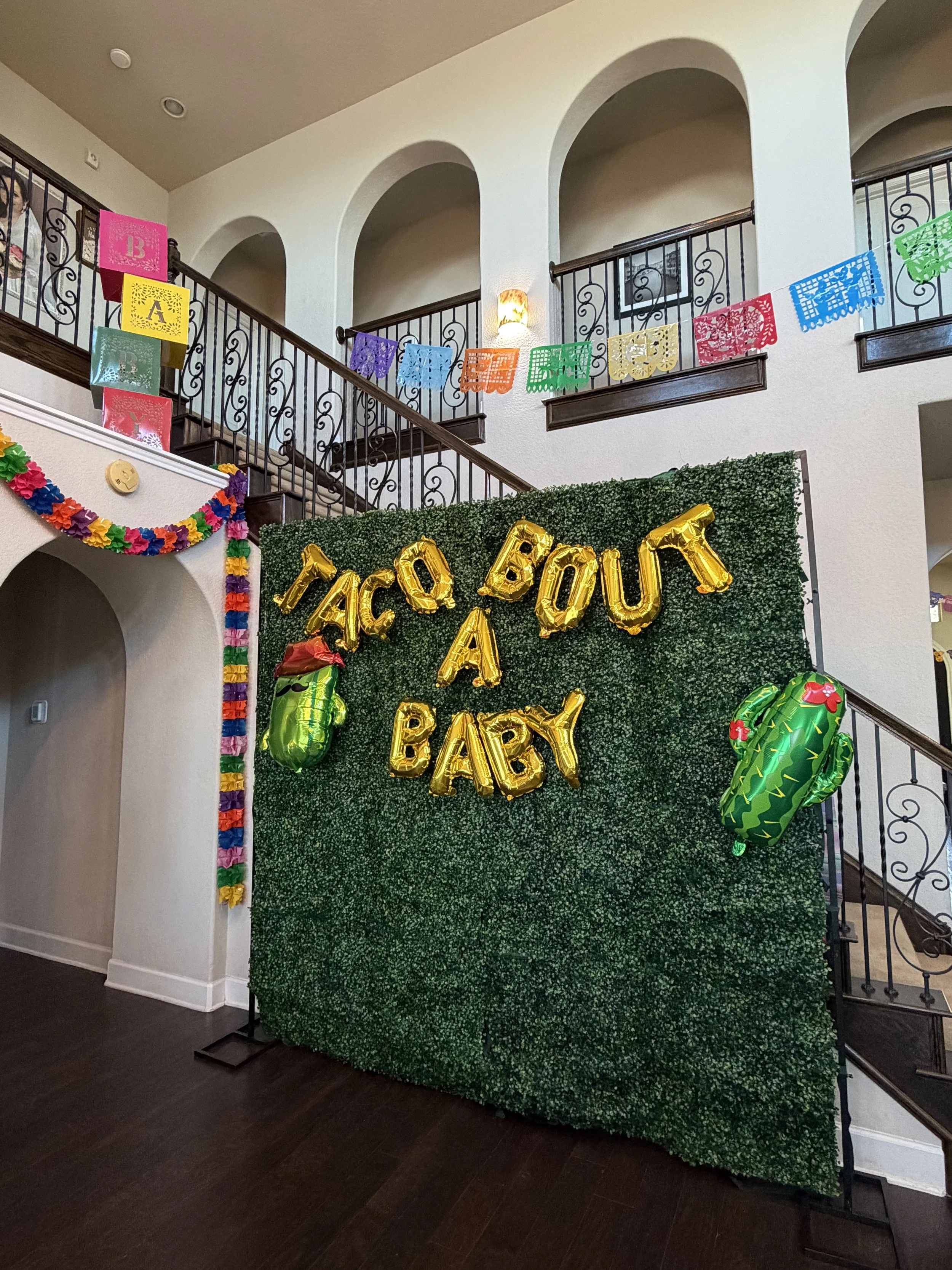 Indoor celebration setup with a green hedge backdrop medium decorated with large gold foil balloons spelling out "TACO BOUT A BABY," two inflatable watermelons, and colorful paper banners and garlands.