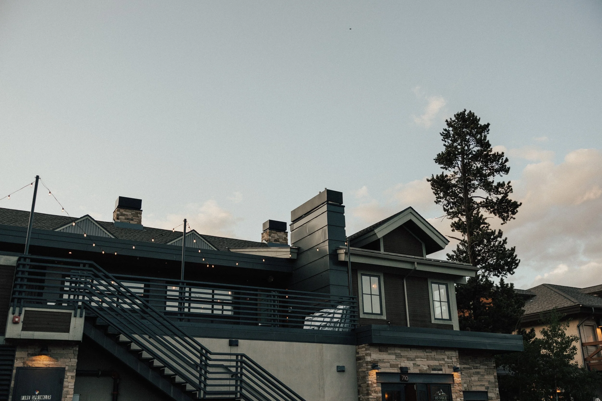 Exterior view of a modern multi-story building with a deck and staircase, string lights hanging, a tall pine tree, and a partly cloudy sky during dusk.