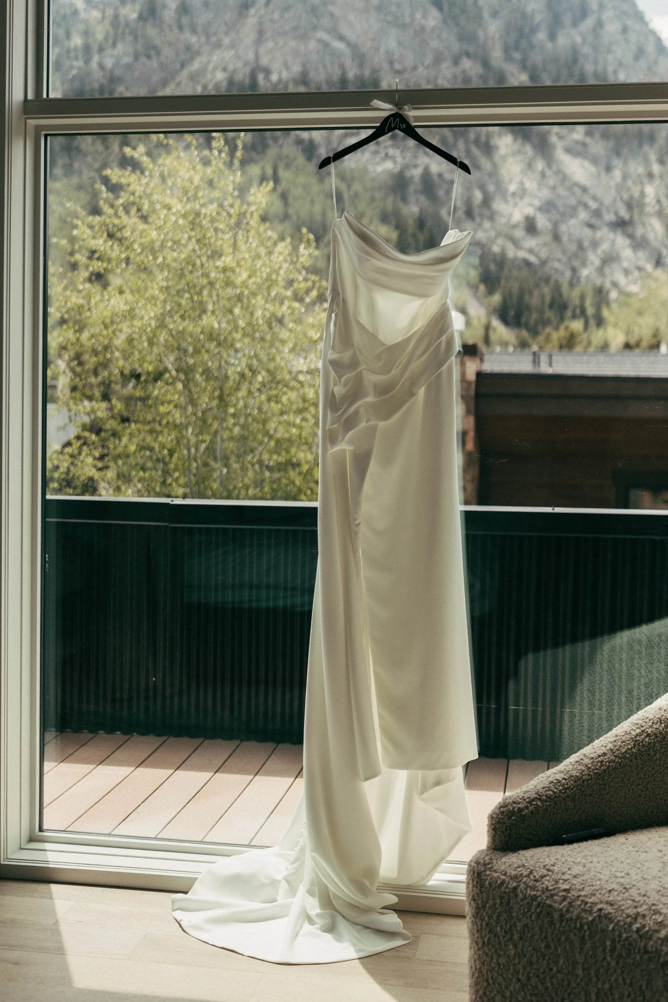 A white wedding dress hanging on a black hanger inside near a glass door, with a mountainous landscape view in the background.